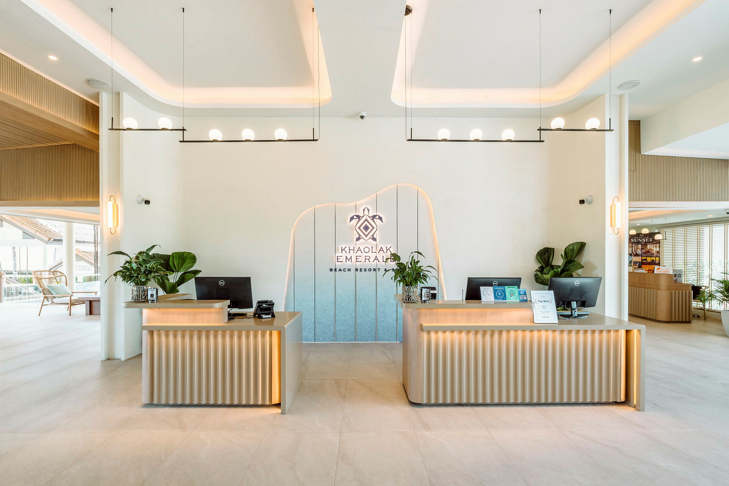 Resepsjonsområde med to moderne pulter dekorert med planter. En logo med teksten 'Khao Lak Emerald Beach Resort' er plassert på veggen. (AI-generert tekst)