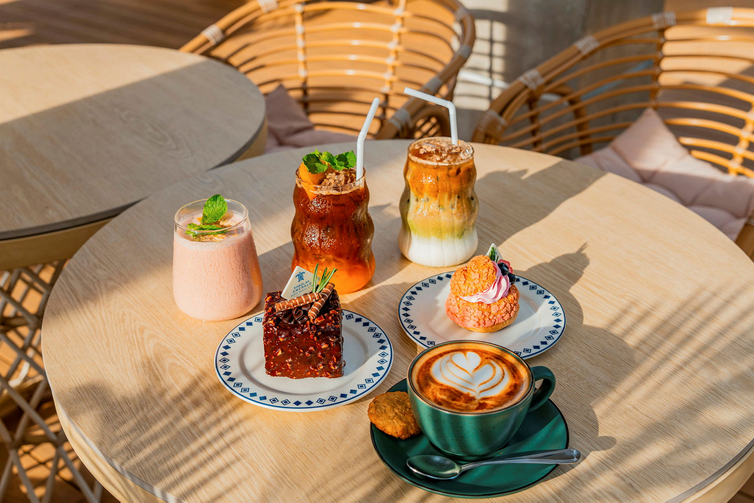 Bildet viser et bord med ulike bakverk og drikker. Det er en kopp kaffe med latte art, en brownie, et rosa bakverk samt noen kalde drikker. (AI-generert tekst)