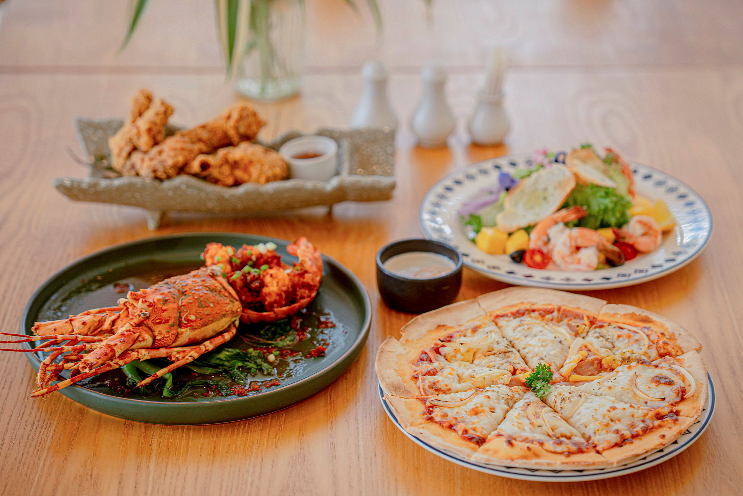 Et matbord med retter som inkluderer en hummer, en tallerken med fritert kylling, en salat med reker og tropiske frukter, samt en pizza. (AI-generert tekst)