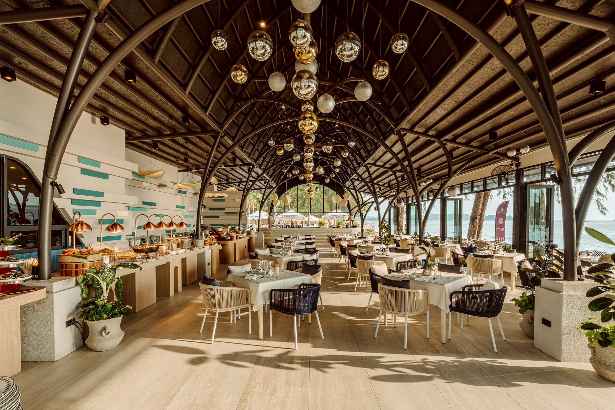 En restaurant med vakker arkitektur og takdekorasjoner, fylt med dekkede bord og en buffébord. Inkluderer utsikt mot havet i bakgrunnen. (AI-generert tekst)