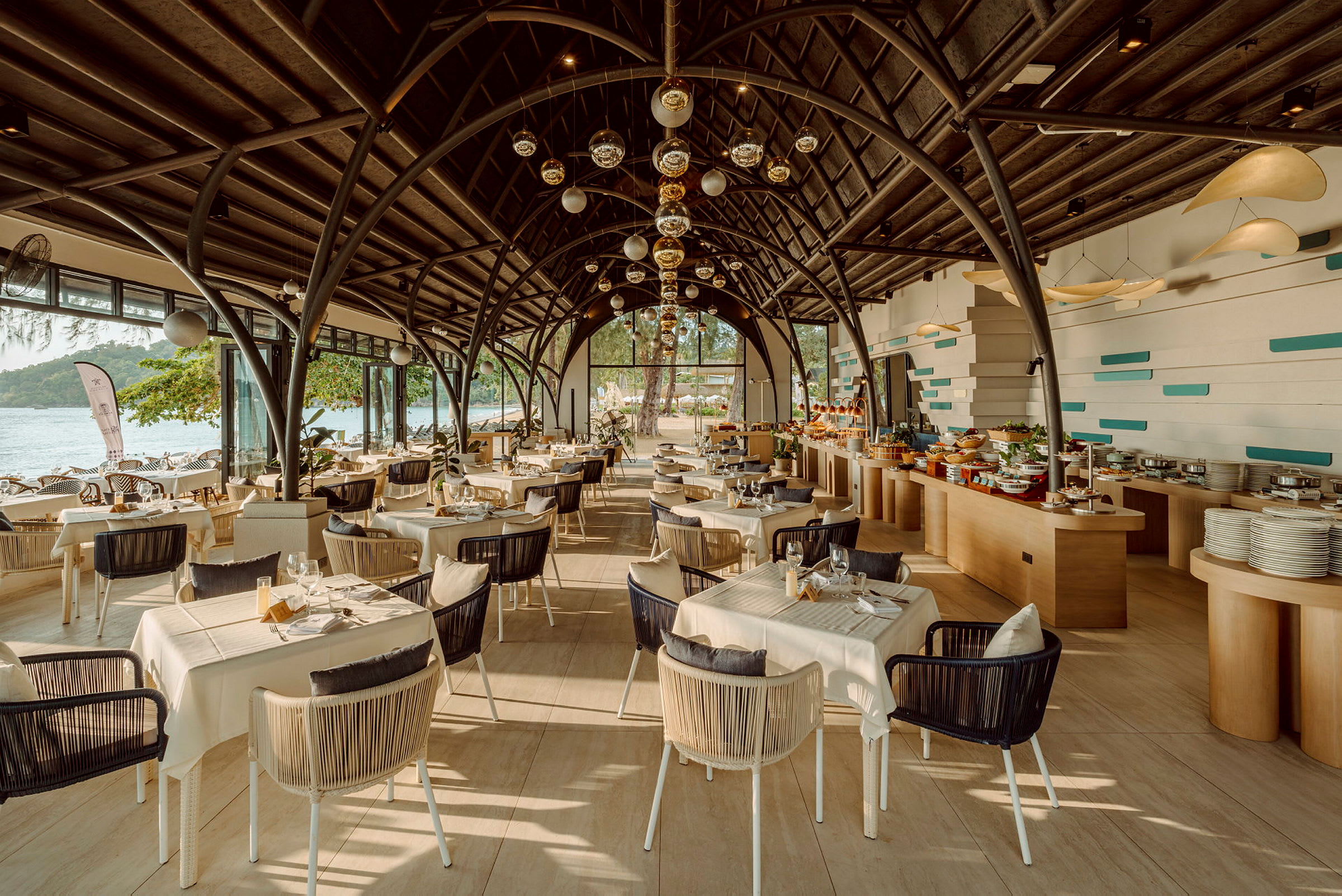 En moderne restaurant med store vinduer som vender mot sjøen. Bord og stoler er pent arrangert under en buet takstruktur, og det er en buffet til høyre. (AI-generert tekst)
