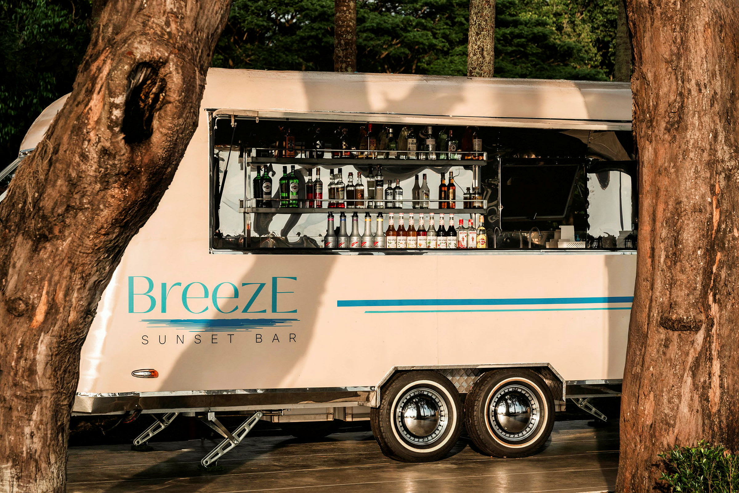 En mobil bardisk med merke 'BreezE Sunset Bar' er plassert mellom trærne, med flasker og drikkevarer synlige i det åpne serviceområdet. (AI-generert tekst)