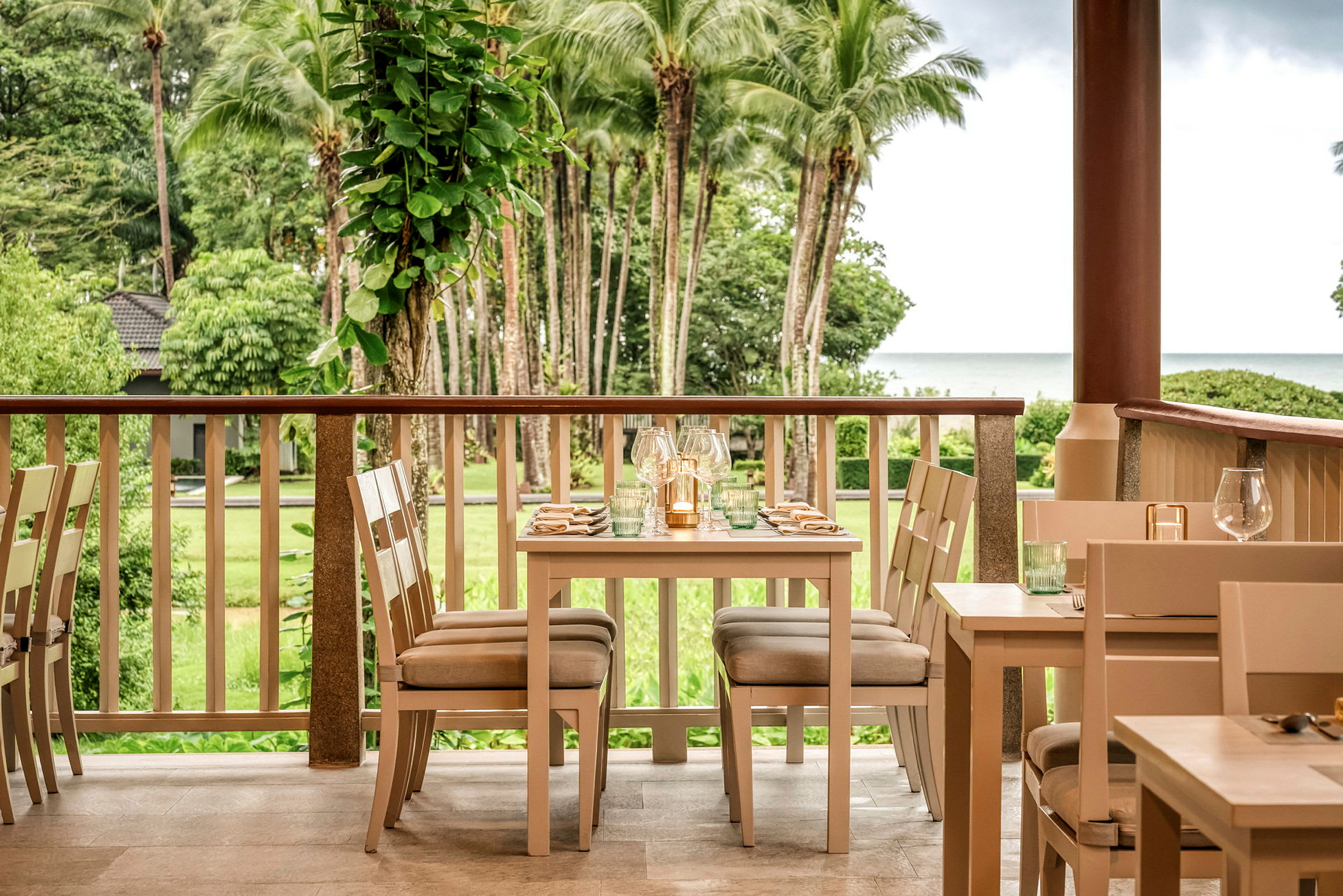 En restaurant med bord og stoler står på en veranda med utsikt over en grønn hage og palmer, samt havet i bakgrunnen. (AI-generert tekst)