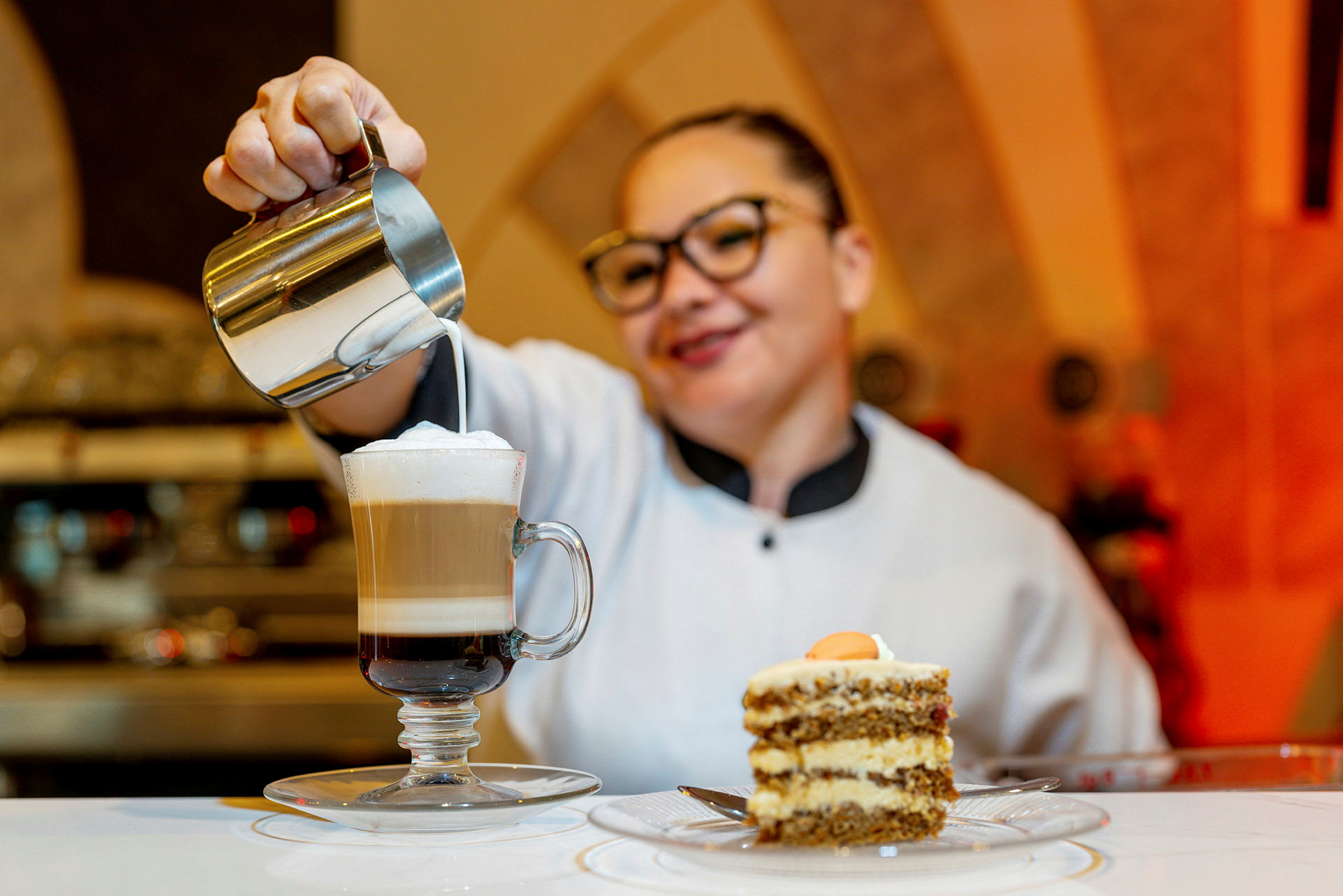 En kvinne heller melk i en kaffe latte foran et kakestykke på et bord. (AI-generert tekst)