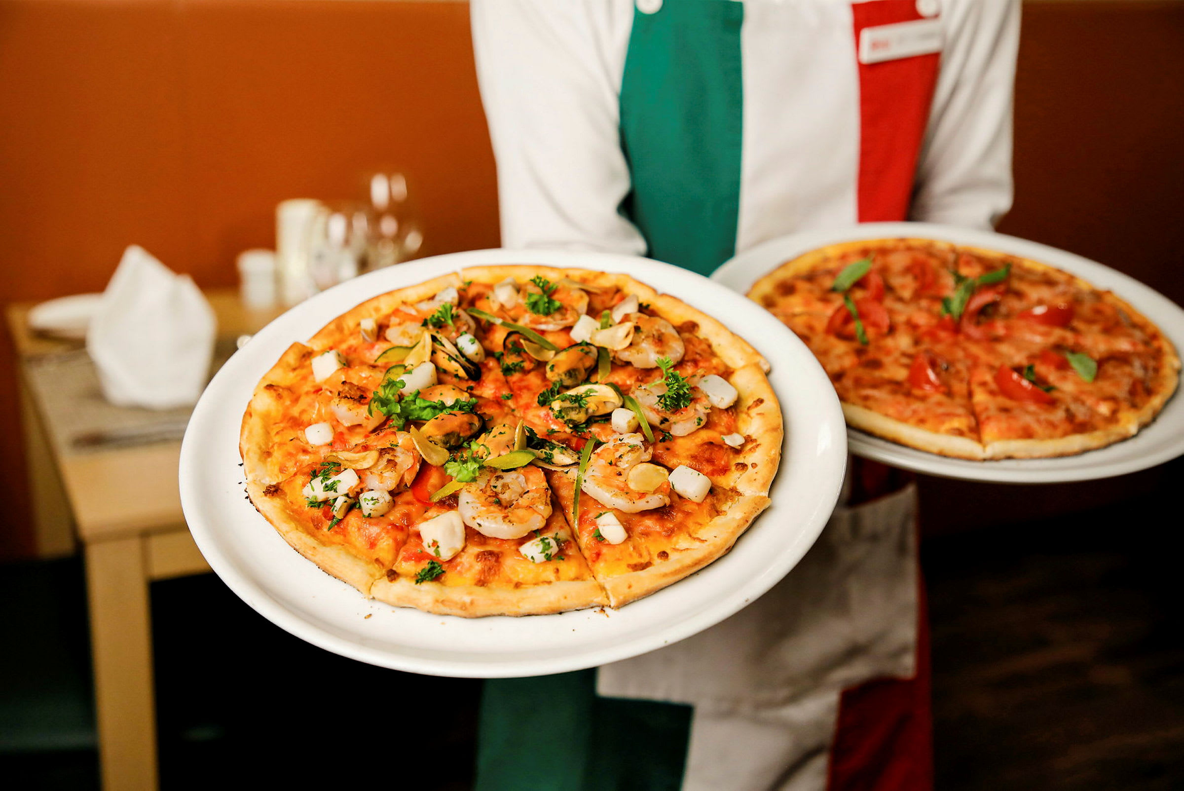 En person holder to tallerkener med pizza, hvorav den ene har reker og urter som topping. (AI-generert tekst)
