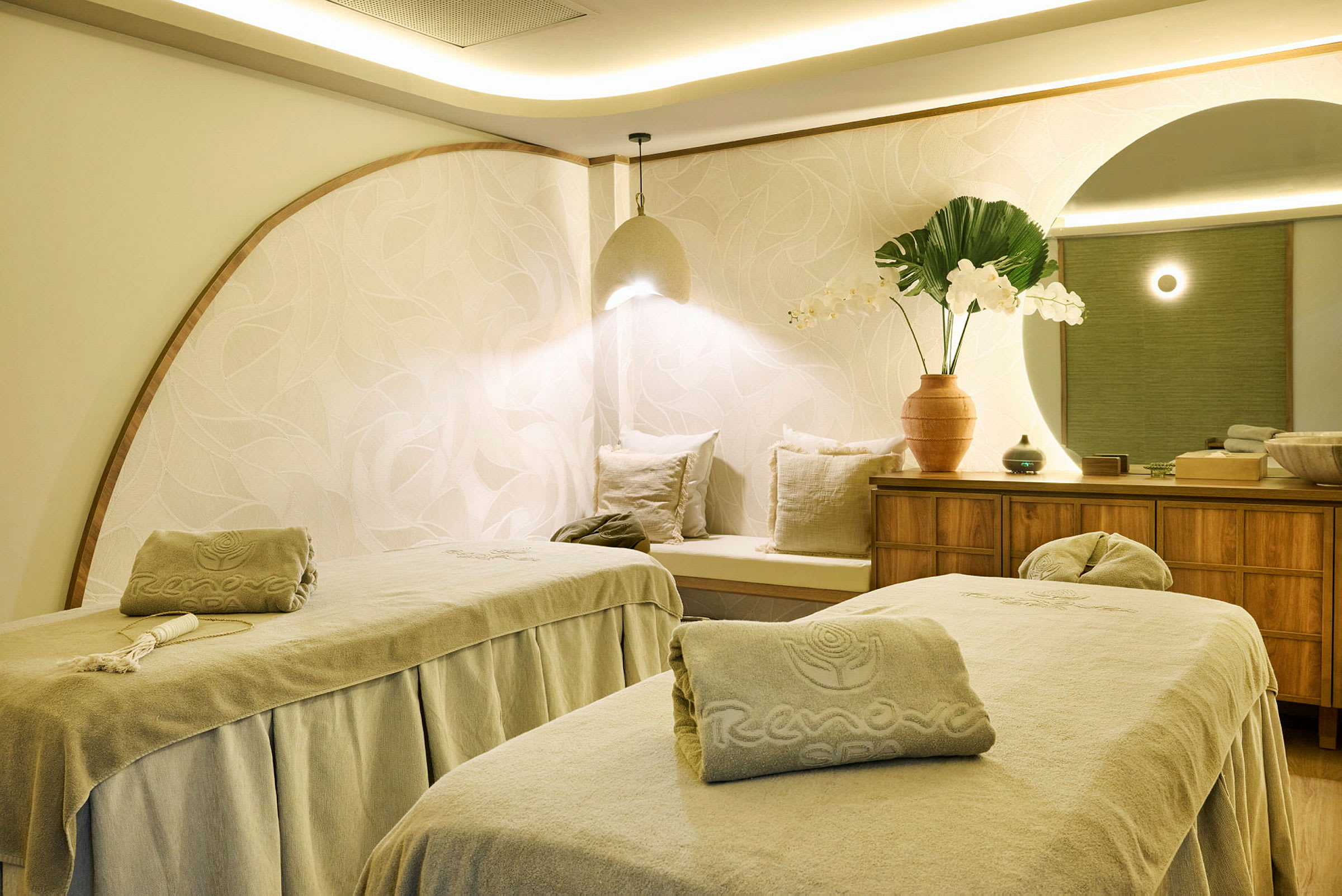 To massasjebord i et godt opplyst og stilfullt innredet spa-rom. På en kommode står dekorative elementer som en vase med blomster. (AI-generert tekst)