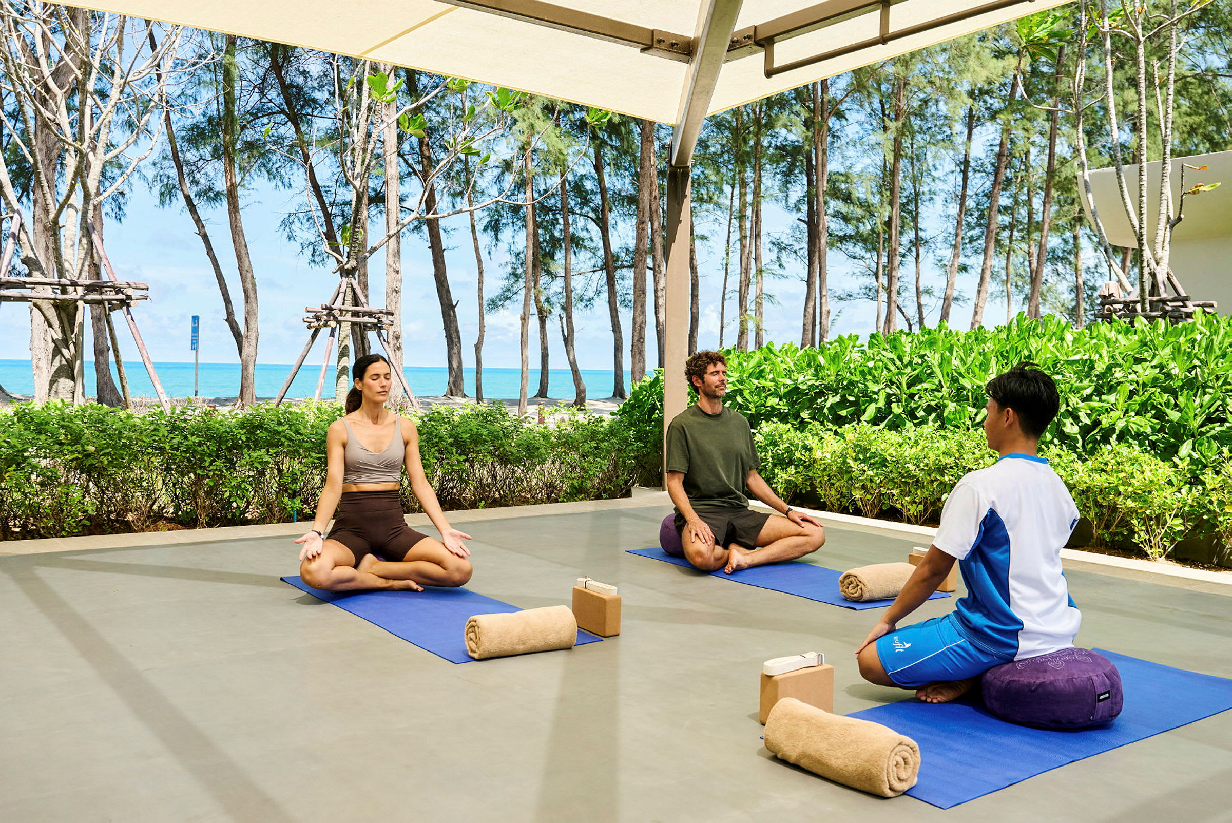 Tre personer sitter på yogamatter utendørs og mediterer. Bak dem er grønne busker, trær og en strand med havet i bakgrunnen. (AI-generert tekst)