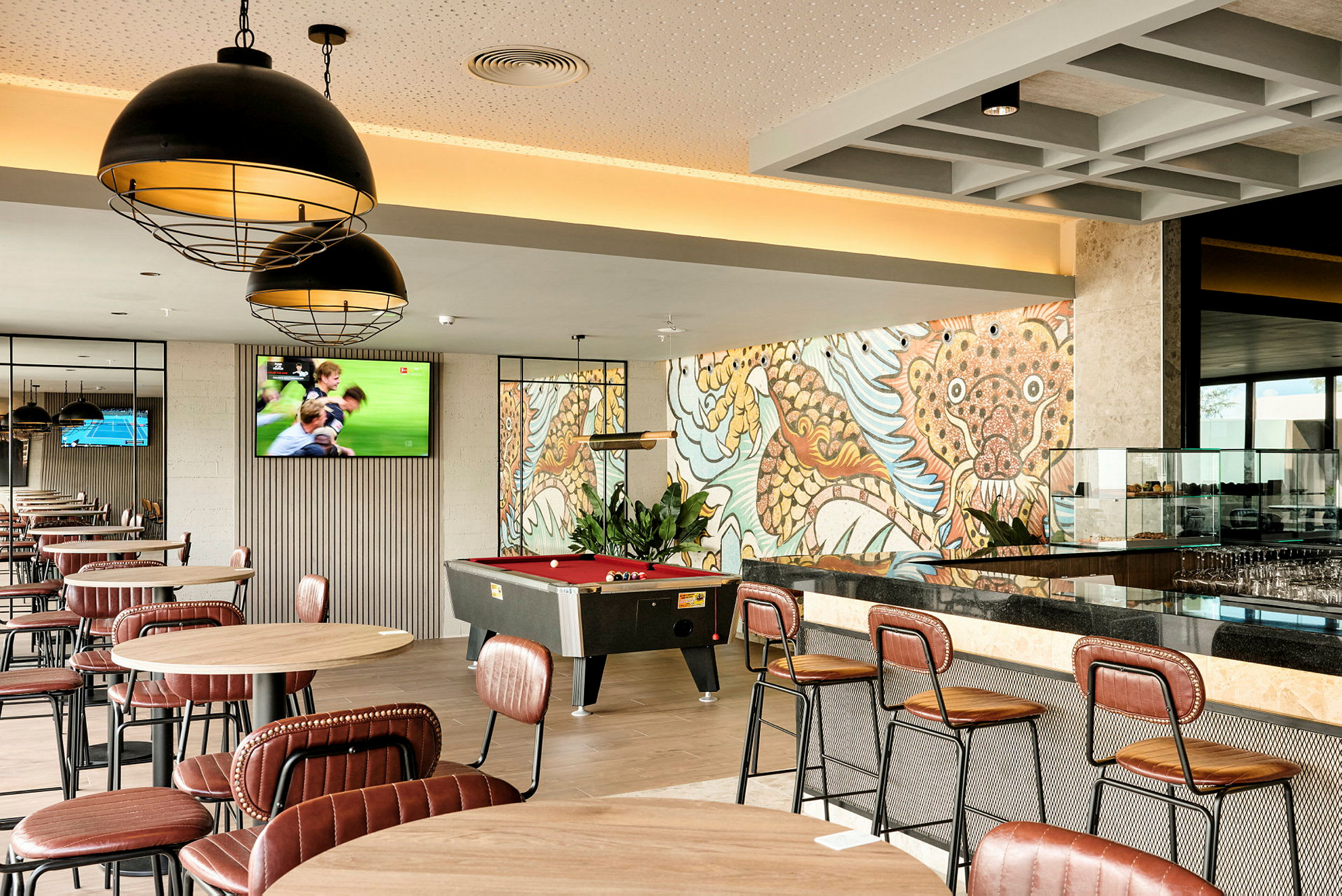 Et kafé eller loungeområde med bord og stoler, et biljardbord og et fargerikt veggmaleri i bakgrunnen. (AI-generert tekst)