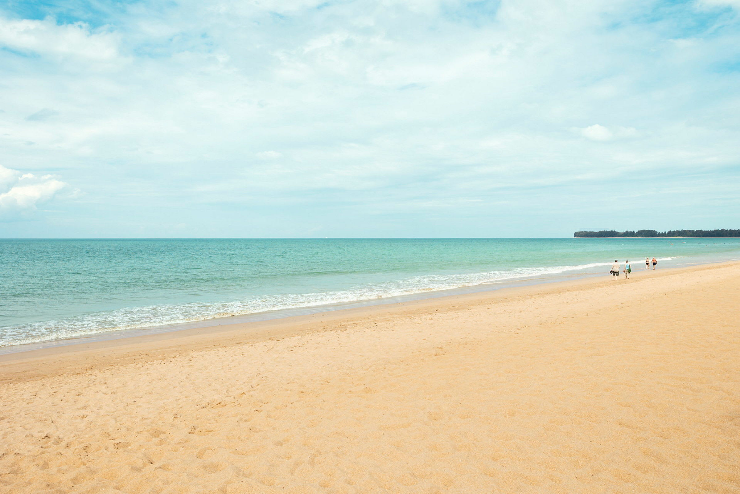 En sandstrand med havet i bakgrunnen og noen personer som går langs stranden. (AI-generert tekst)