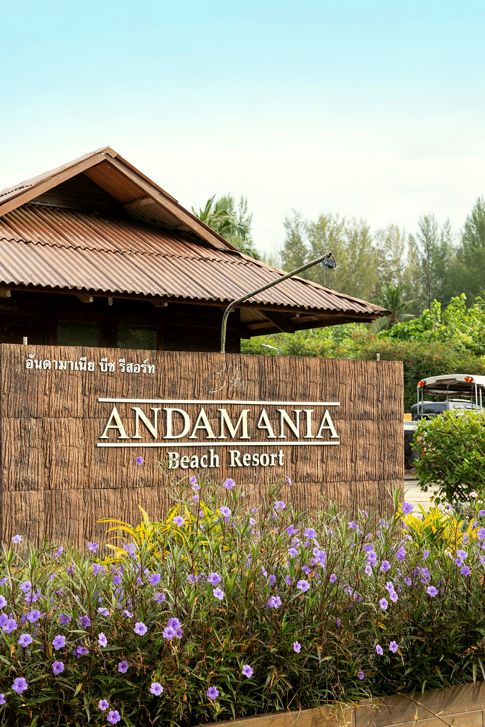 Inngangen til Andamania Beach Resort, med et skilt omgitt av lilla blomster og en bygning med et blikktak i bakgrunnen. (AI-generert tekst)