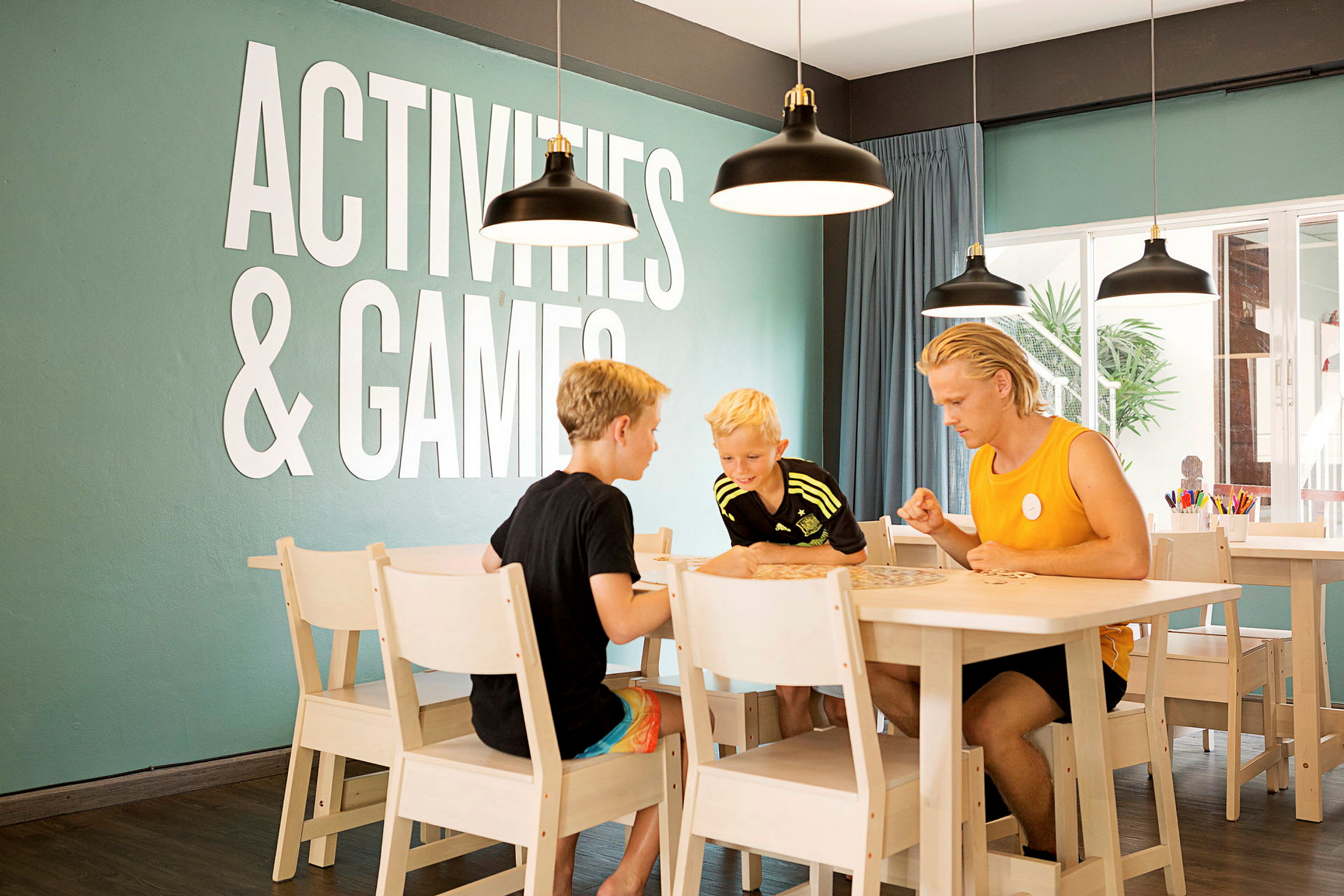Personer spiller et bordspill ved et bord i et rom med teksten "Activities & Games" på veggen. (AI-generert tekst)