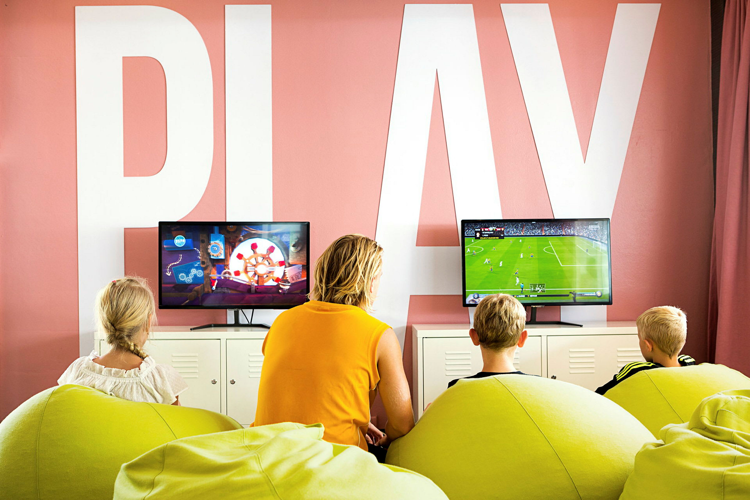 Tre barn og en voksen sitter på grønne sakkosekker og spiller TV-spill foran to skjermer i et rom med teksten 'PLAY' på veggen. (AI-generert tekst)