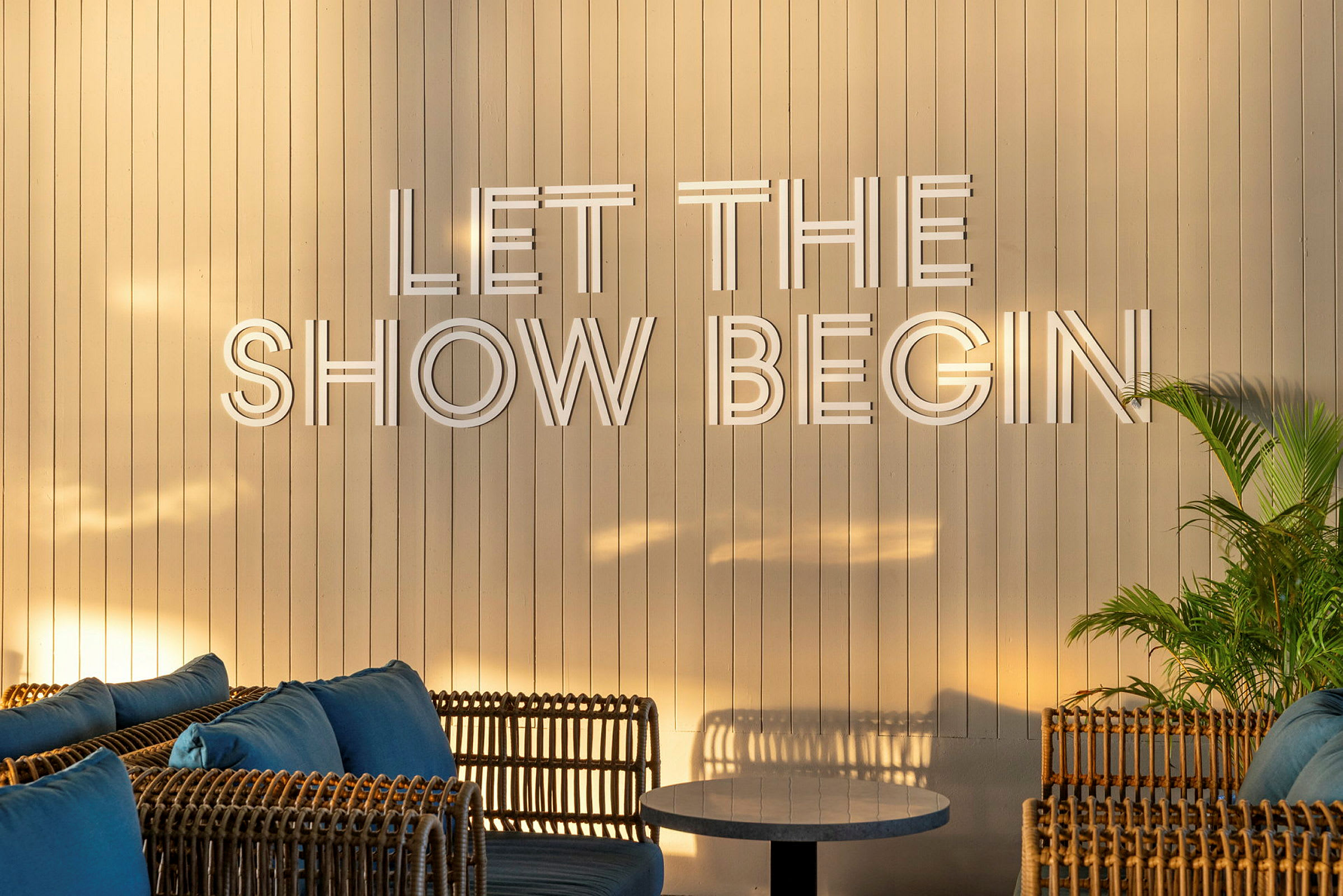 En lys trepanelvegg med teksten "Let the show begin", omgitt av rottingmøbler og grønne planter. (AI-generert tekst)