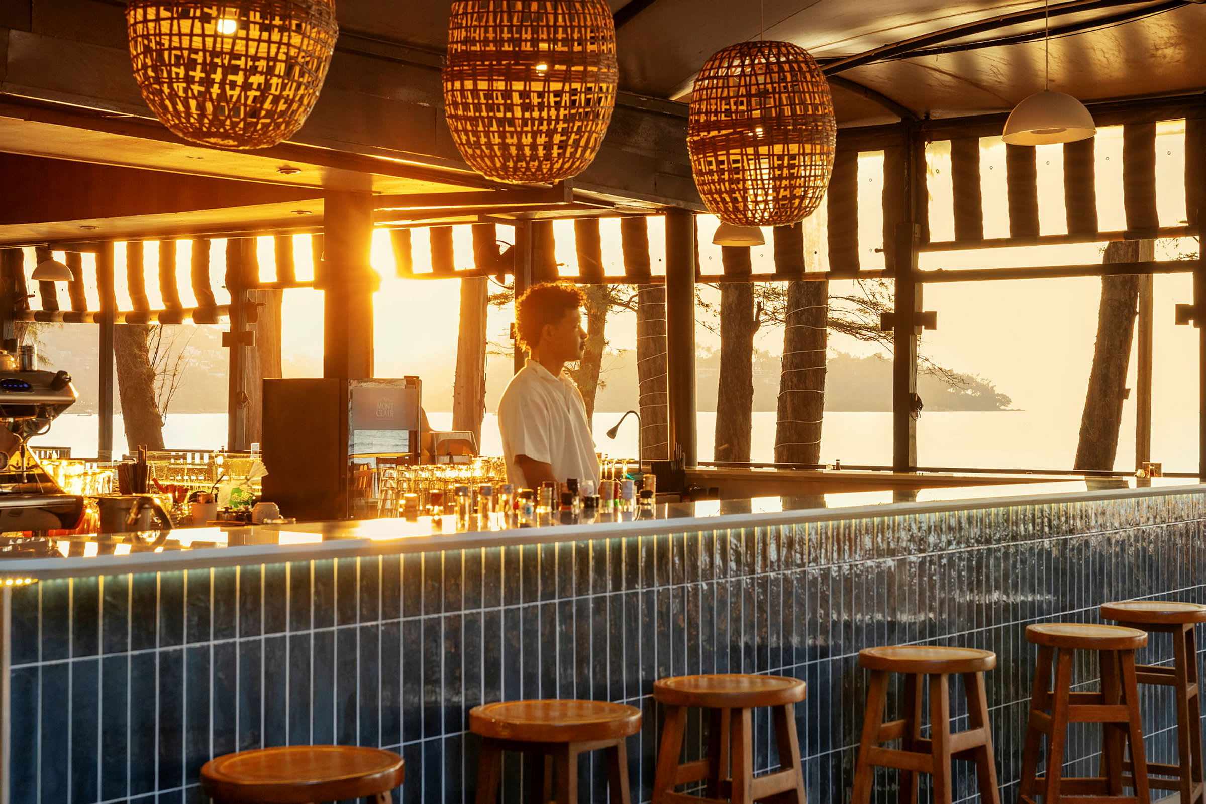 En elegant bar belyst av varmt sollys gjennom vinduene, med runde trekrakker foran en lang bardisk. (AI-generert tekst)