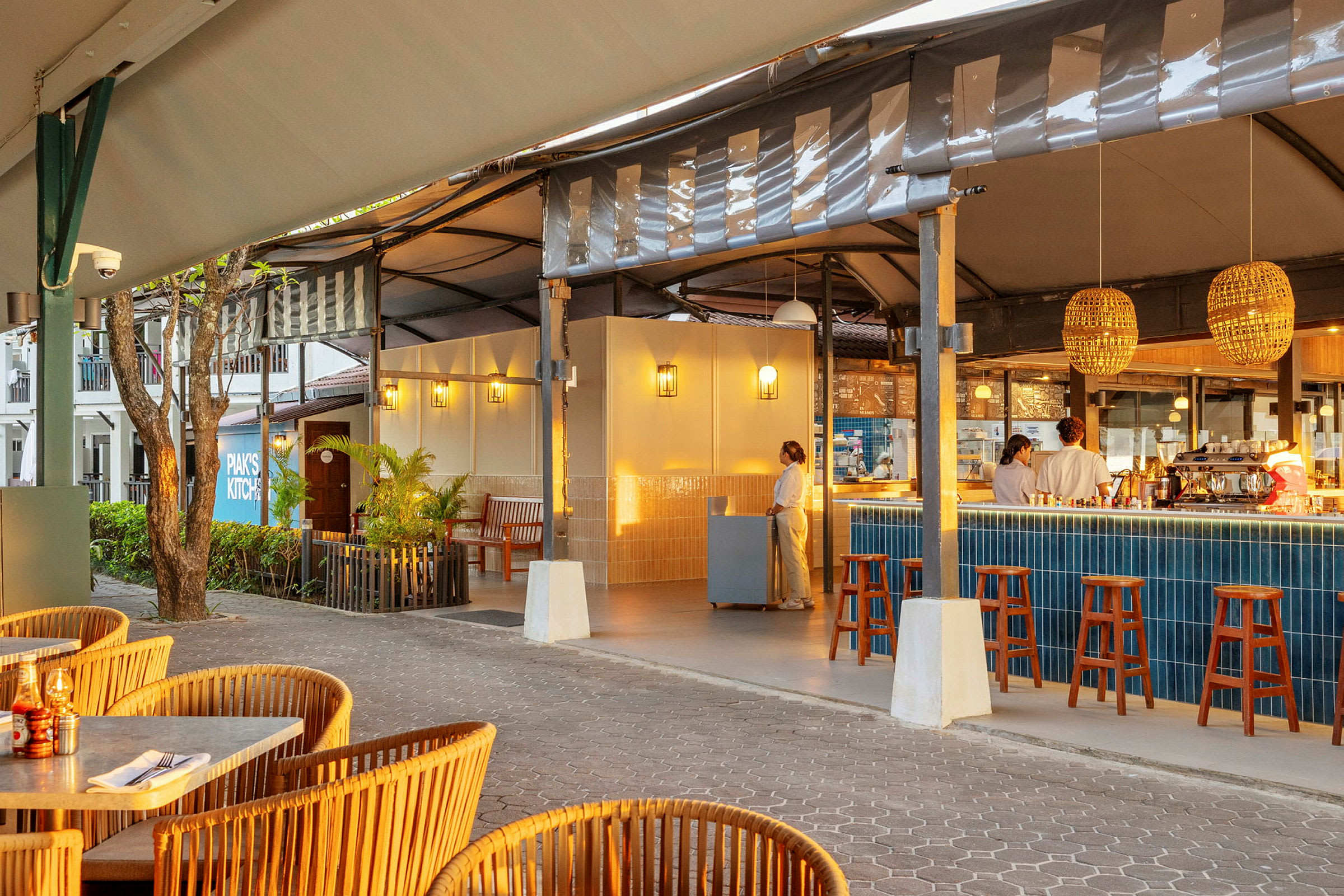 En åpen restaurant med flettede stoler og et barområde med høye stoler og blå fliser. Personale er synlige ved disken. (AI-generert tekst)