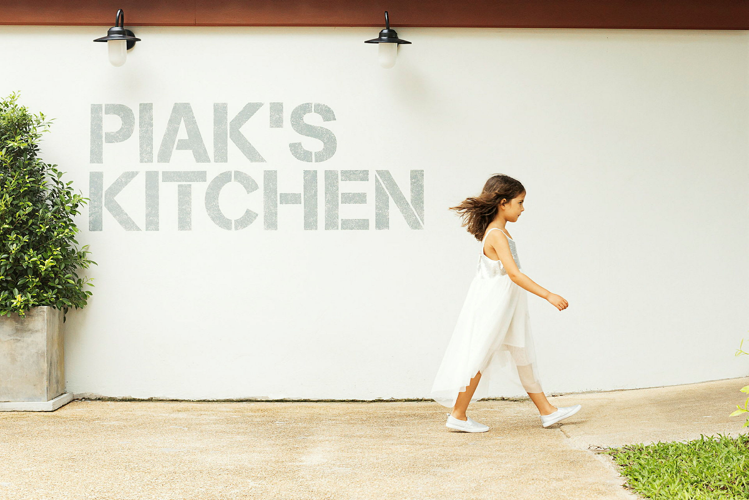 En person går forbi en hvit vegg med teksten 'PIAK'S KITCHEN' og en lampe samt en potteplante synlig. (AI-generert tekst)
