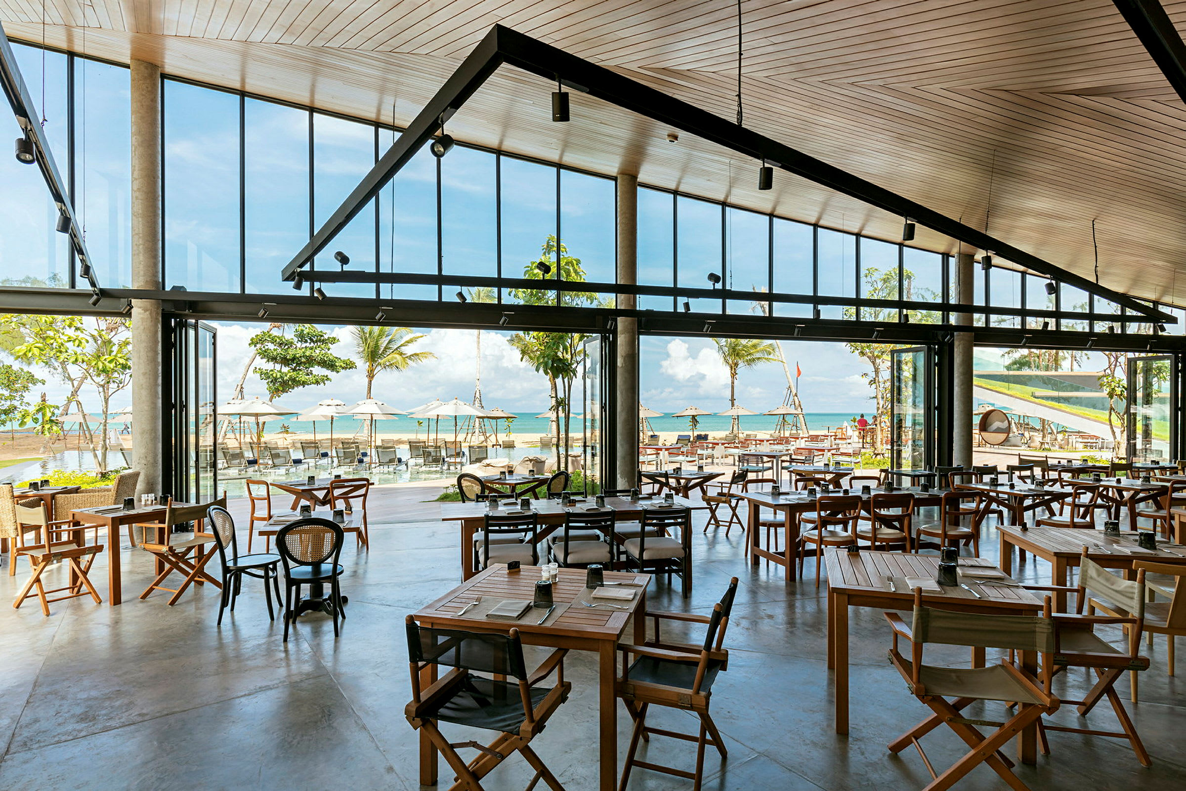 Et åpent og lyst restaurantområde med store vinduer som vender mot stranden og havet med palmer og parasoller. (AI-generert tekst)