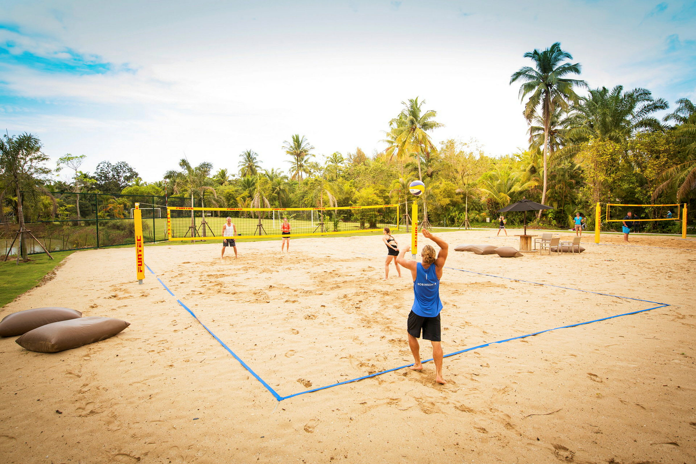 Flere personer spiller sandvolleyball på en sandbane omgitt av palmer og grønne omgivelser. (AI-generert tekst)