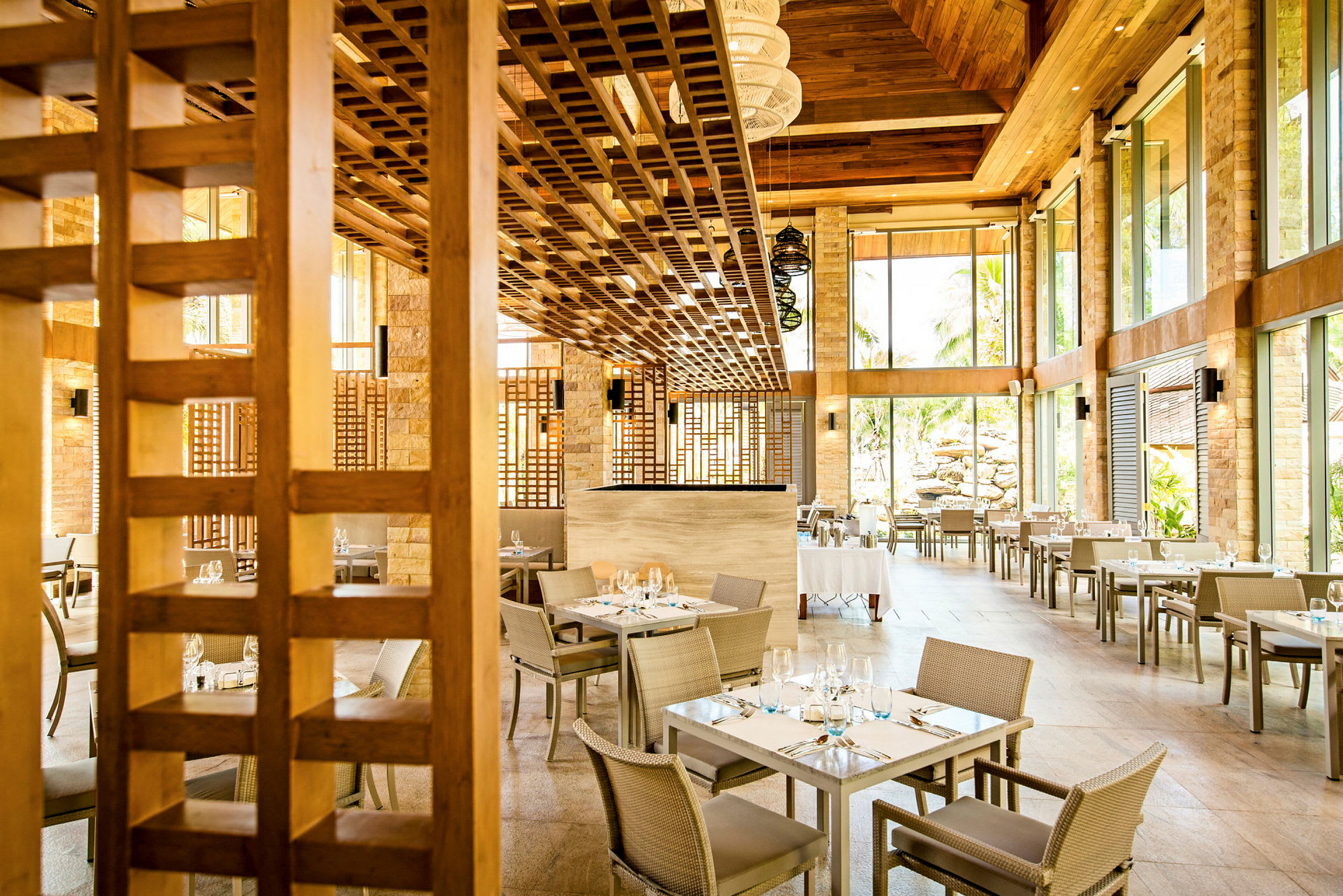 En lys restaurant med sitteplasser ved firkantede bord og store glassvegger som slipper inn naturlig lys. Taket er laget av tre. (AI-generert tekst)