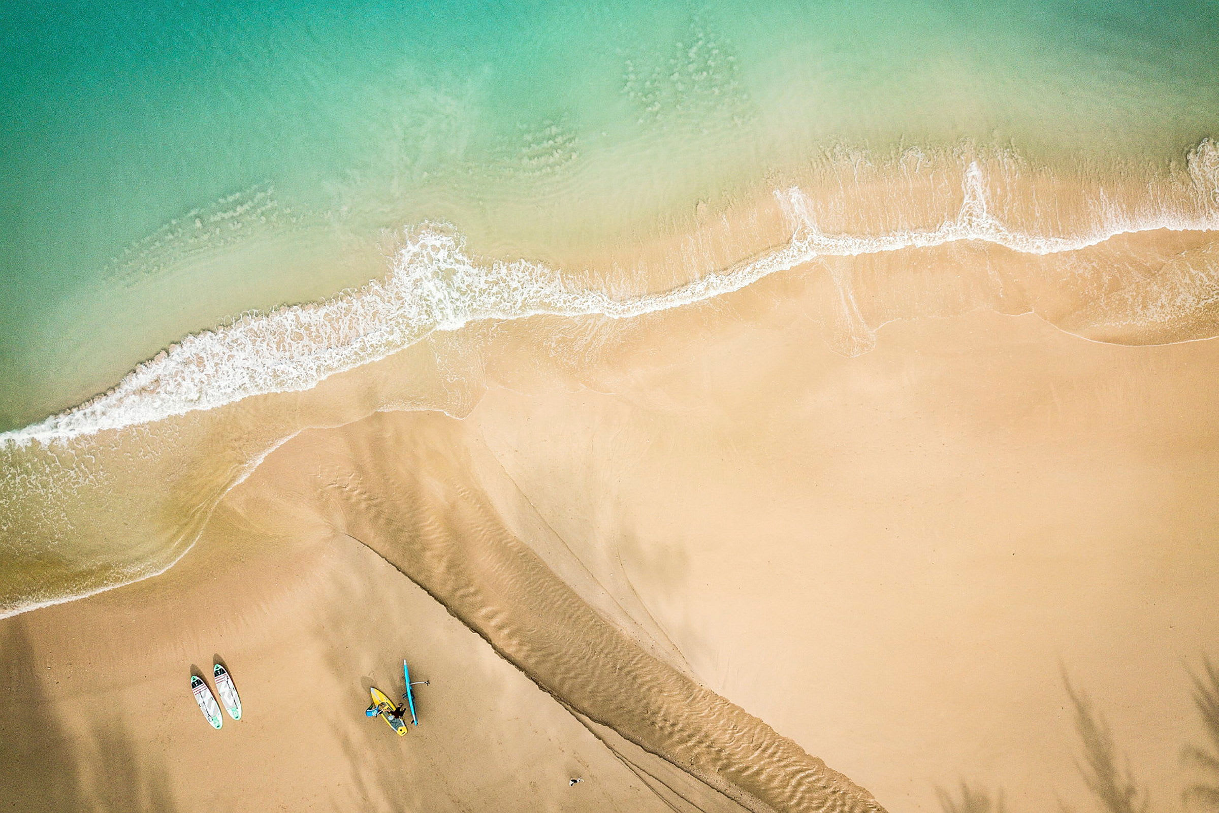 Flyfotografi av en sandstrand hvor bølger møter stranden. To paddleboard og en kajakk er synlige på sanden. (AI-generert tekst)