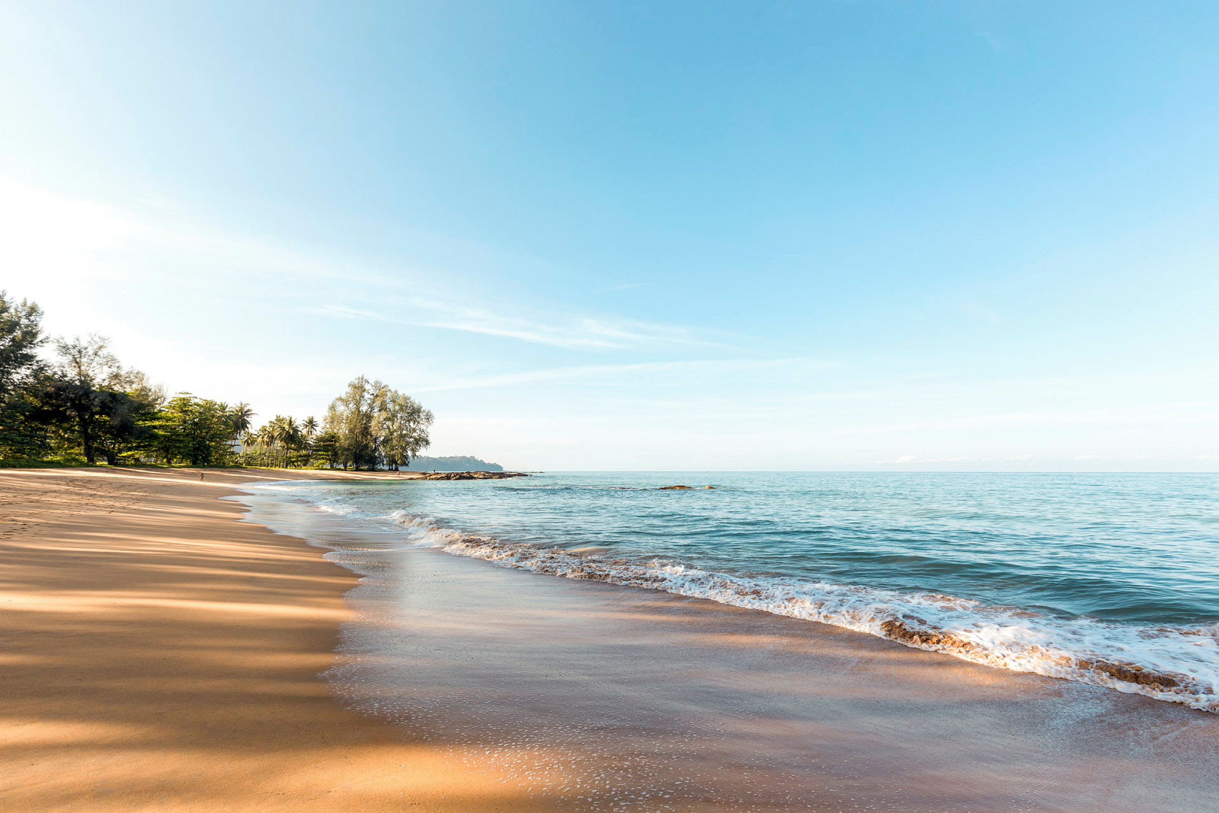En sandstrand med havet og strandkant i forgrunnen, trær er synlige i bakgrunnen. (AI-generert tekst)