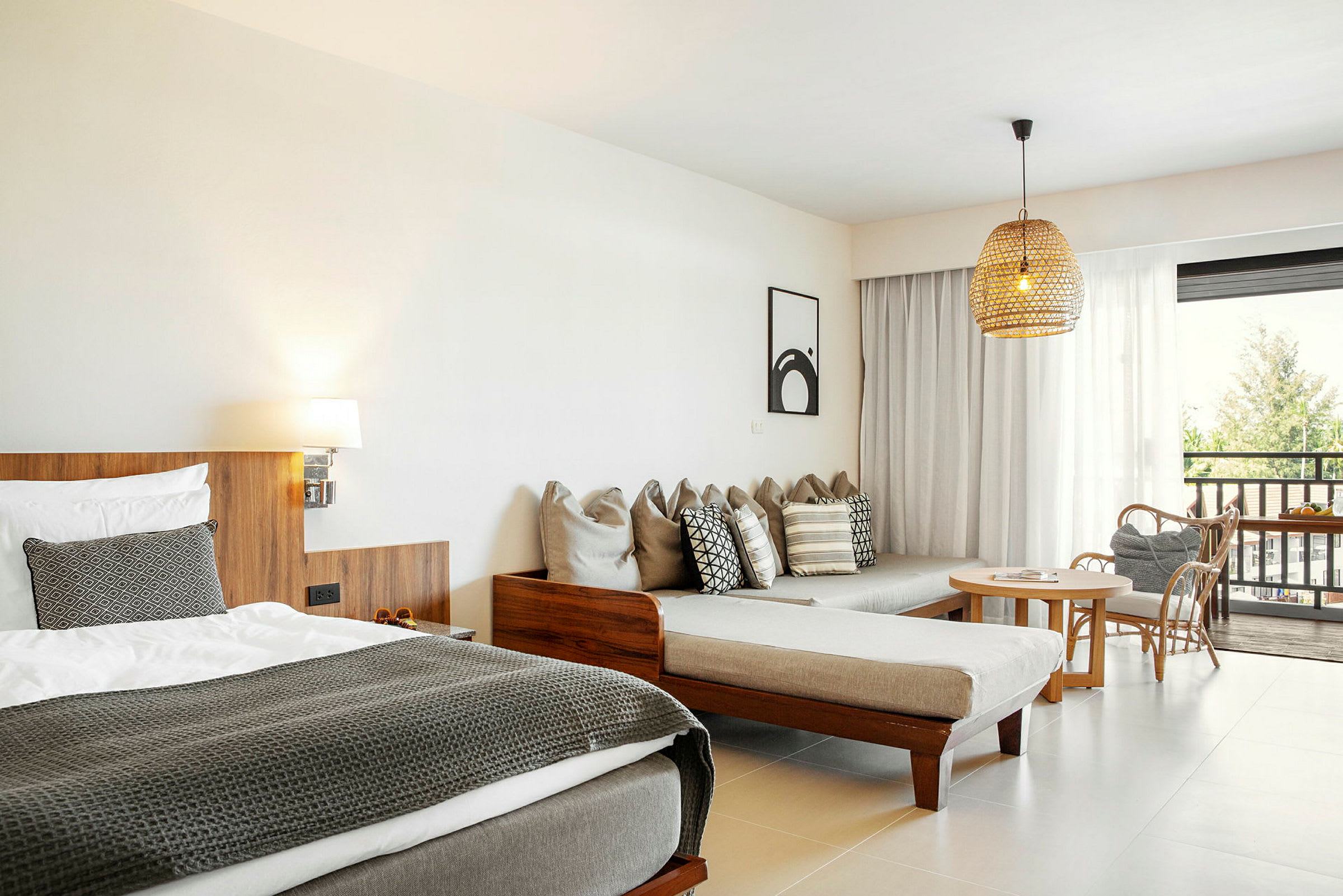 Et hotellrom med en seng, en sofa med puter, et bord med stoler og utsikt utendørs gjennom glassdører. (AI-generert tekst)