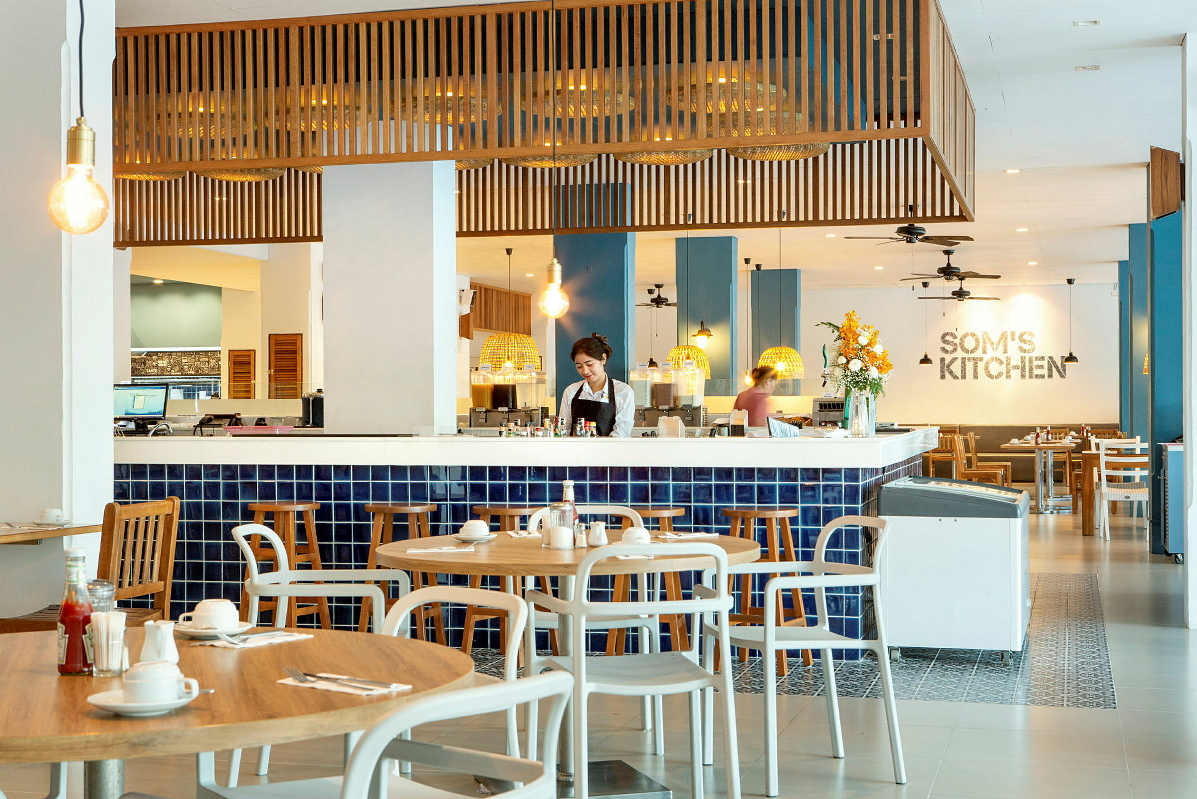Bilde av en restaurant med moderne interiør og blå fliser, der trebord og stoler er arrangert rundt omkring. (AI-generert tekst)