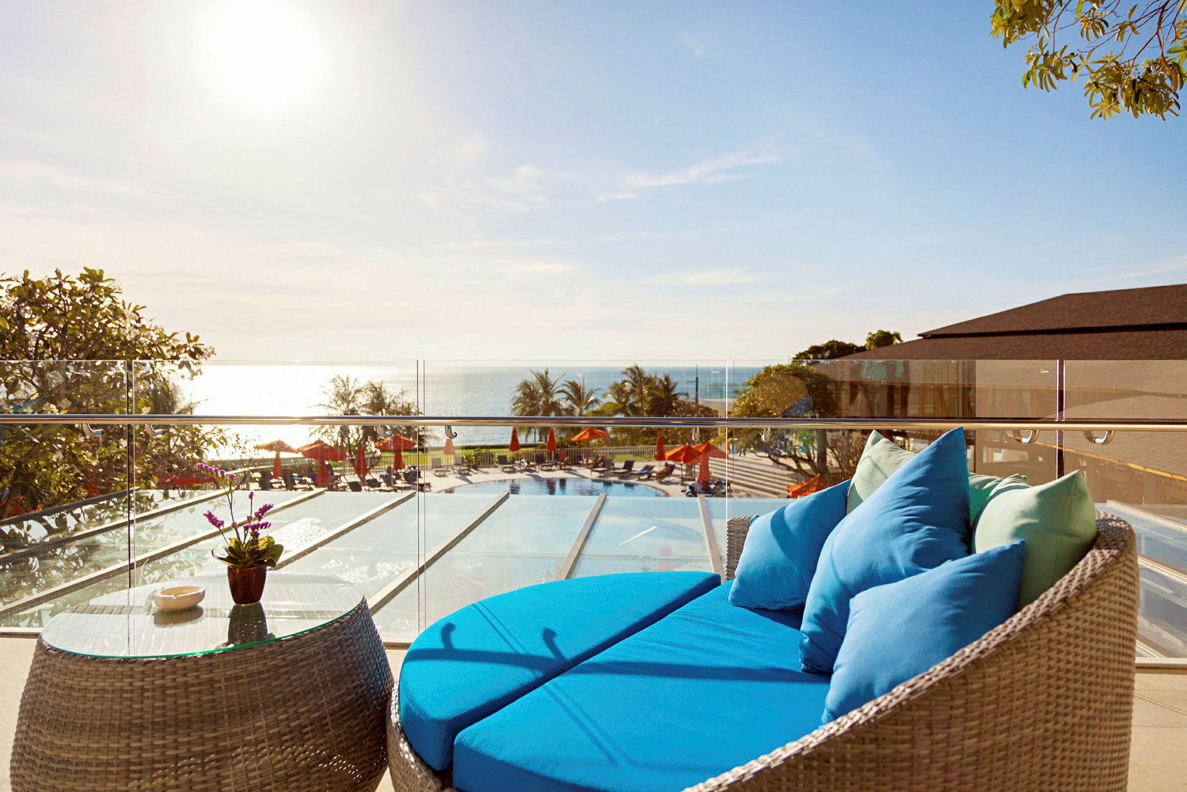 En terrasse med en blå sofa og et glassbord, med utsikt over et basseng, havet og solsenger under parasoller. (AI-generert tekst)