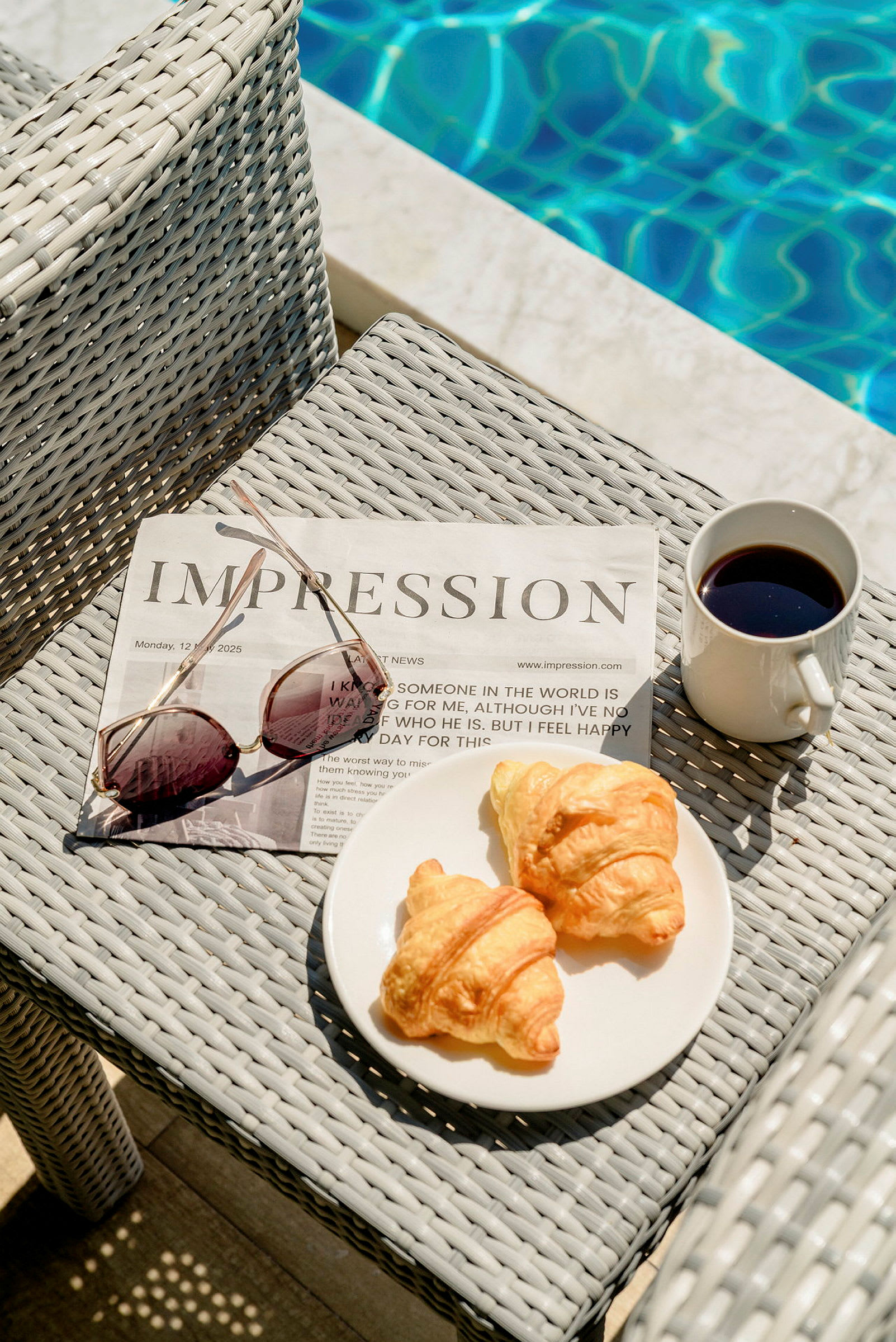 En avis og solbriller på en bordflate ved siden av en tallerken med to croissanter og en kopp kaffe, med et basseng i bakgrunnen. (AI-generert tekst)