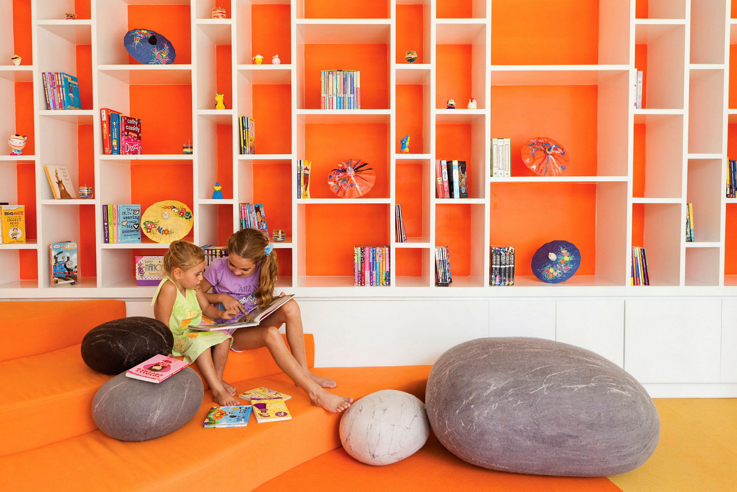 To personer sitter og leser en bok i et fargerikt bibliotek med oransje bakgrunn og hyller fylt med bøker og dekorasjoner. (AI-generert tekst)