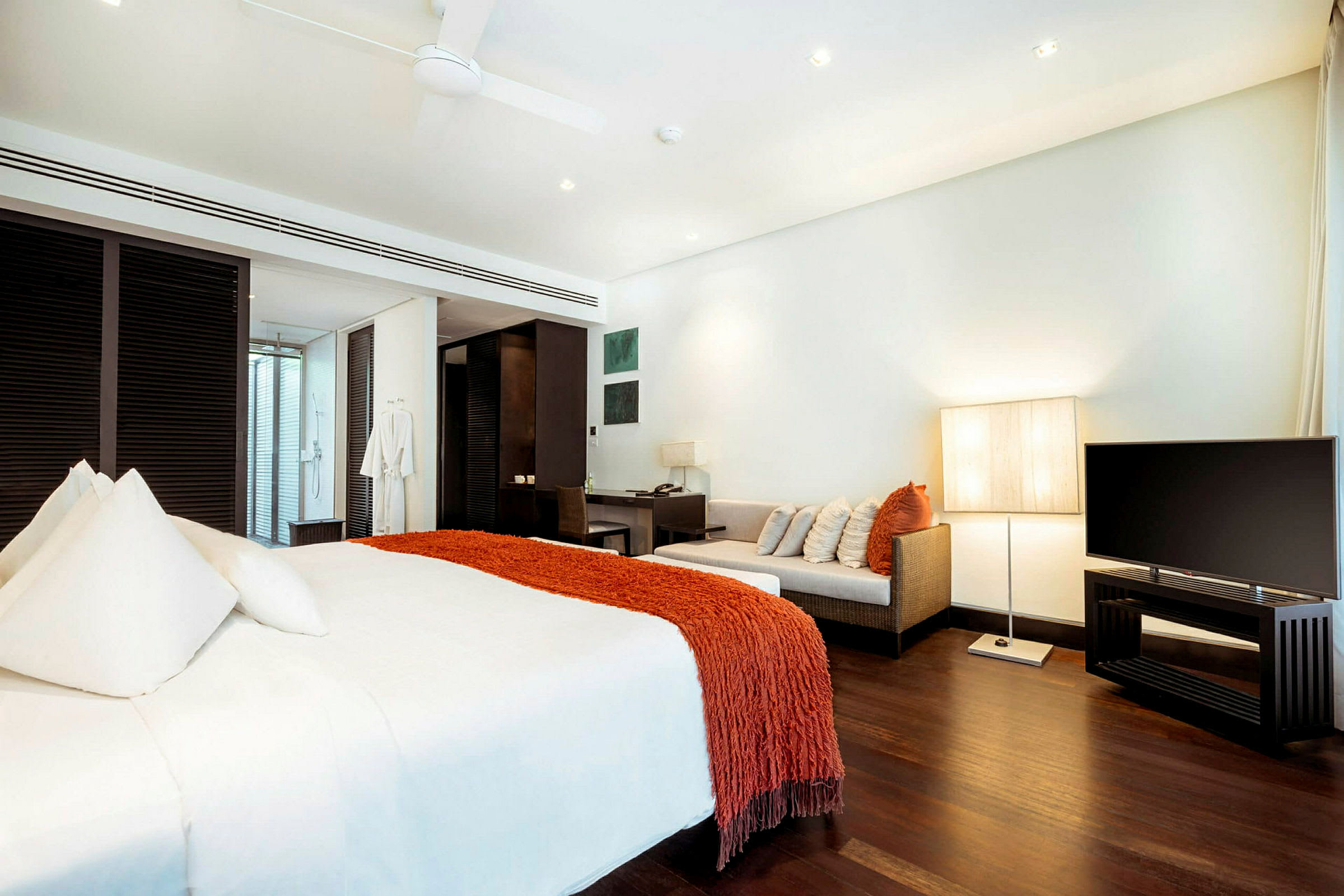 Et moderne hotellrom med en stor seng med hvitt sengetøy og et oransje teppe, en sofa med puter, en skrivebordshjørne og en TV. Rommet har mørkt tregulv og er godt opplyst. (AI-generert tekst)
