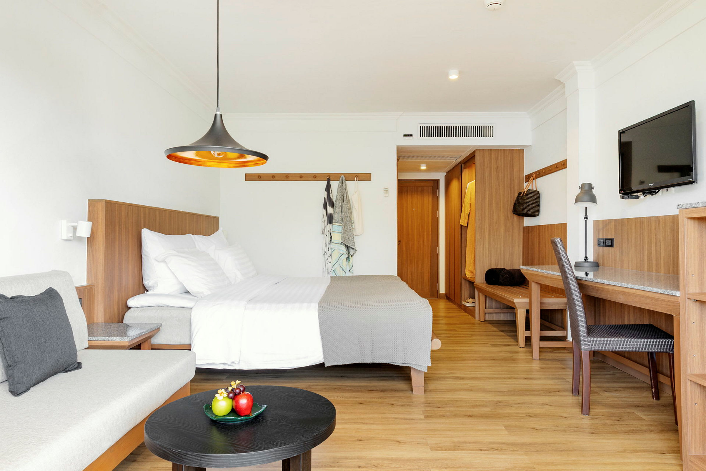 Et moderne hotellrom med seng, sofa, skrivebord og en TV, innredet i lyse farger og tredetaljer. (AI-generert tekst)