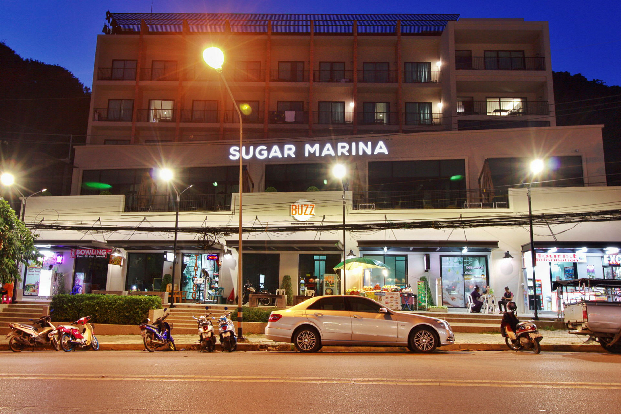 Bygning med hotellnavnet Sugar Marina belyst på fasaden om natten, med motorsykler og biler parkert ved gaten. (AI-generert tekst)