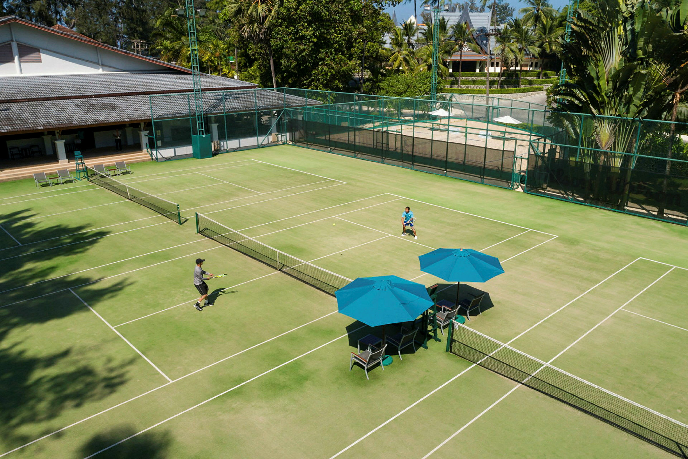 En grønn tennisbane med to personer som spiller tennis. Det er blå parasoller og stoler ved siden av banen. (AI-generert tekst)