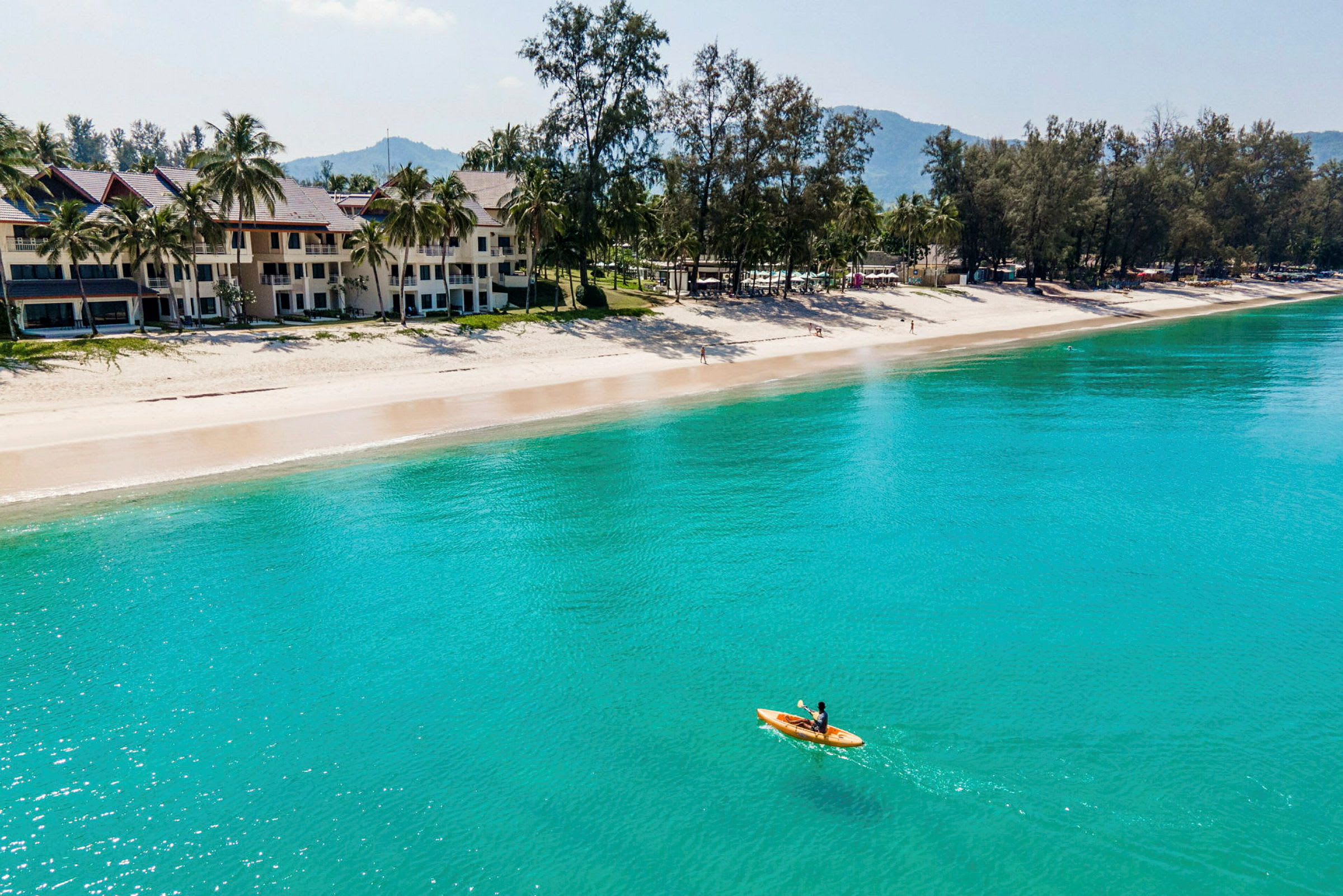 En strand med grønne palmer og bygninger i bakgrunnen, samt en person som padler kajakk på det klare vannet. (AI-generert tekst)