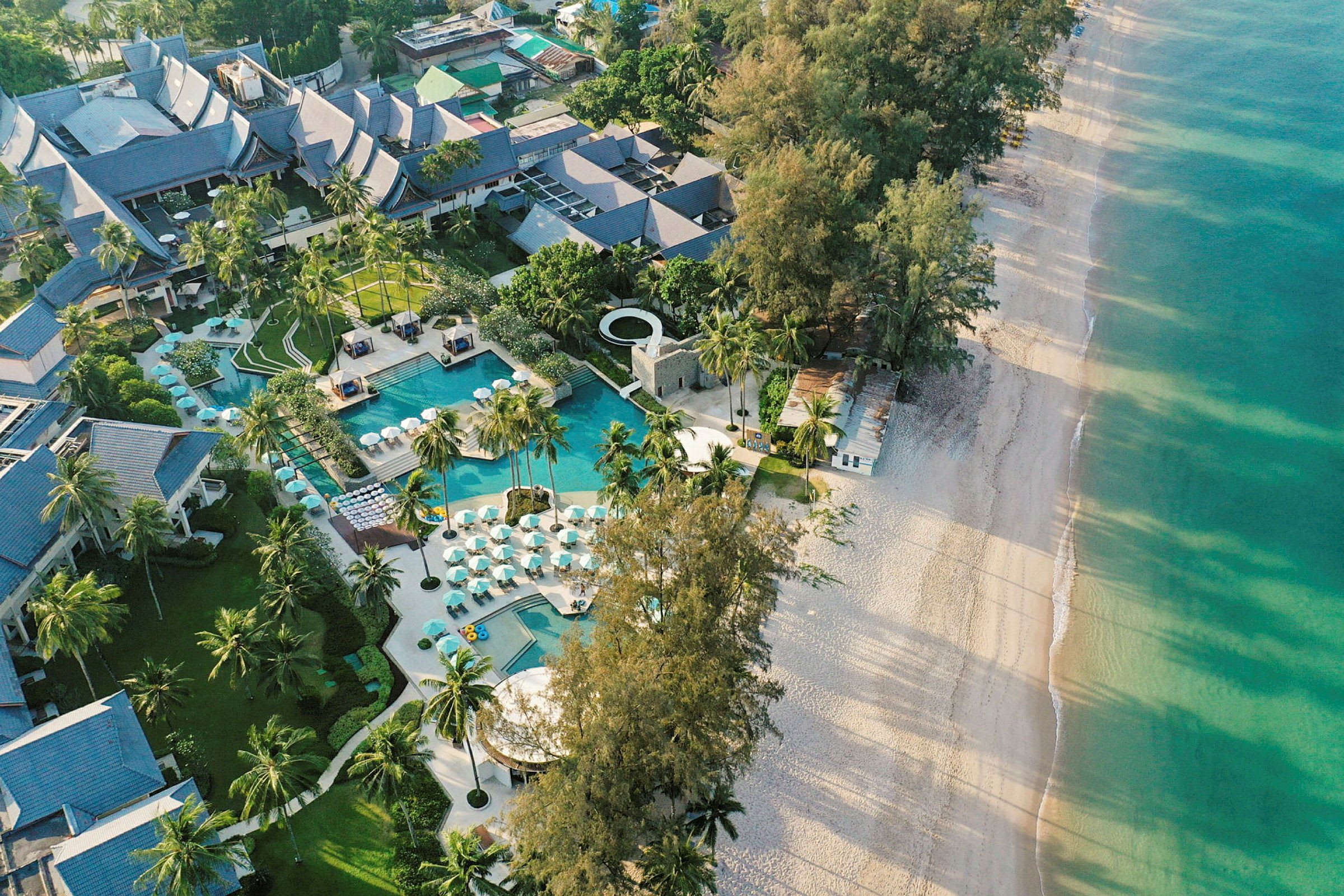 Flyfoto av en resort med basseng og omgitt av palmer ved en sandstrand. (AI-generert tekst)