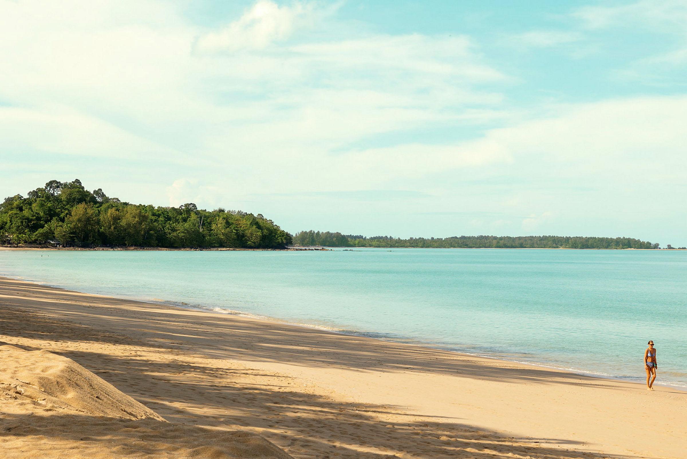 Strand med gyllen sand, rolig blå sjø og frodig skog i bakgrunnen. En person går langs vannkanten. (AI-generert tekst)