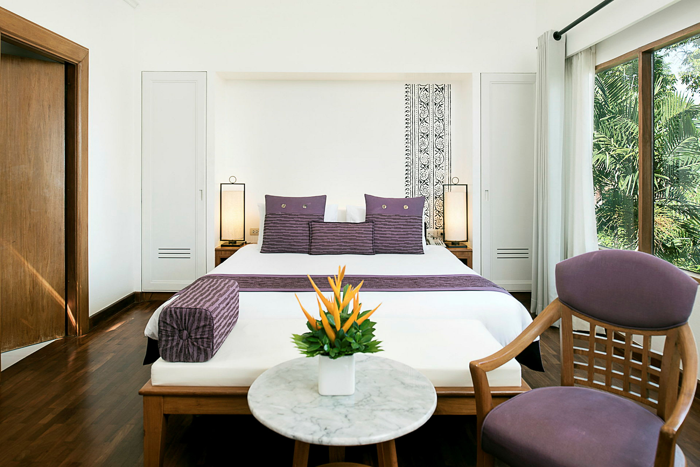 Et hotellrom med en dobbeltseng og lilla dekorative puter, et rundt marmorbord med en blomsterpotte, en stol og et vindu som gir utsikt til grønne områder utenfor. (AI-generert tekst)