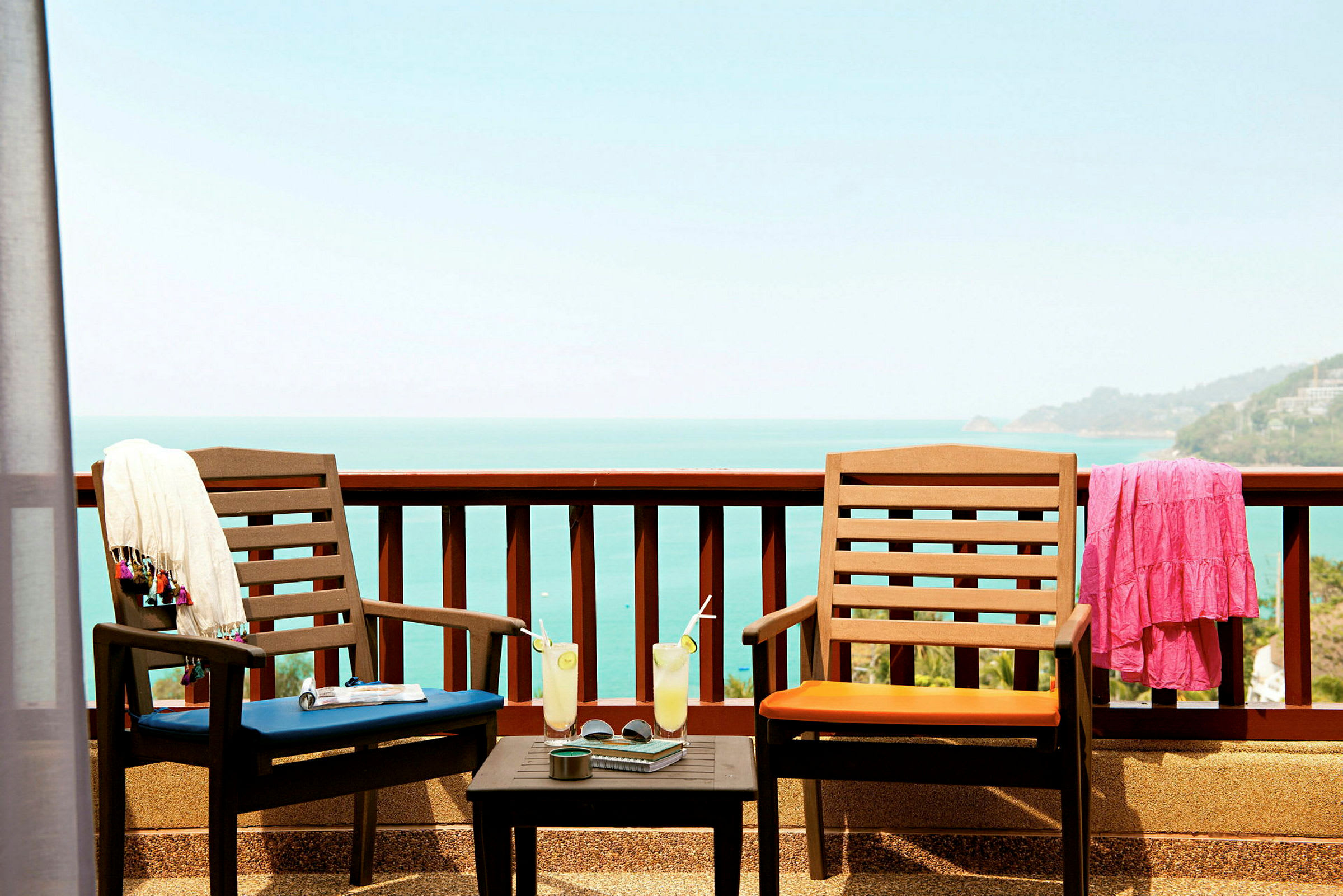 To utestoler med puter står på en terrasse med utsikt over havet. På bordet mellom stolene står glass med drikke og en kopp. Et skjerf og en kjole henger på stolryggene. (AI-generert tekst)