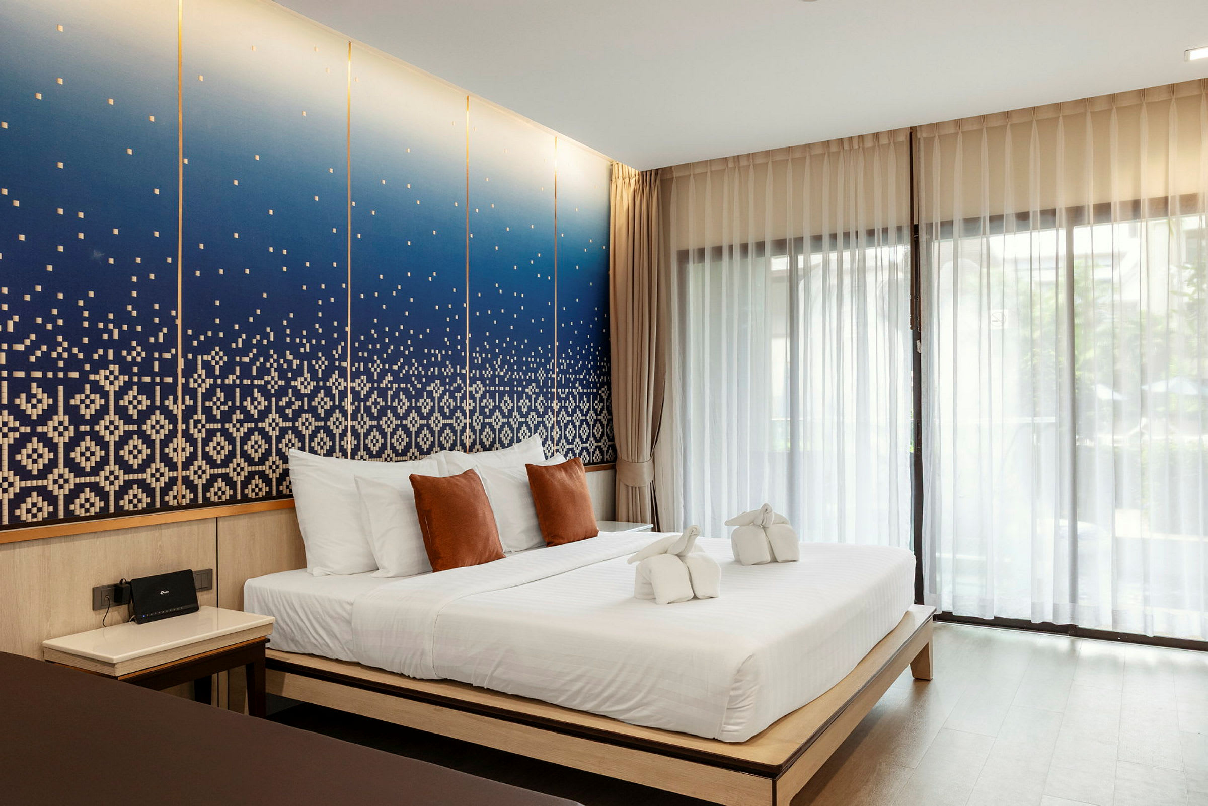 Et hotellrom med en dobbelseng, hvite og brune puter samt dekorative håndklær på sengen. Bakgrunnen har en blå kunstnerisk veggdesign. (AI-generert tekst)