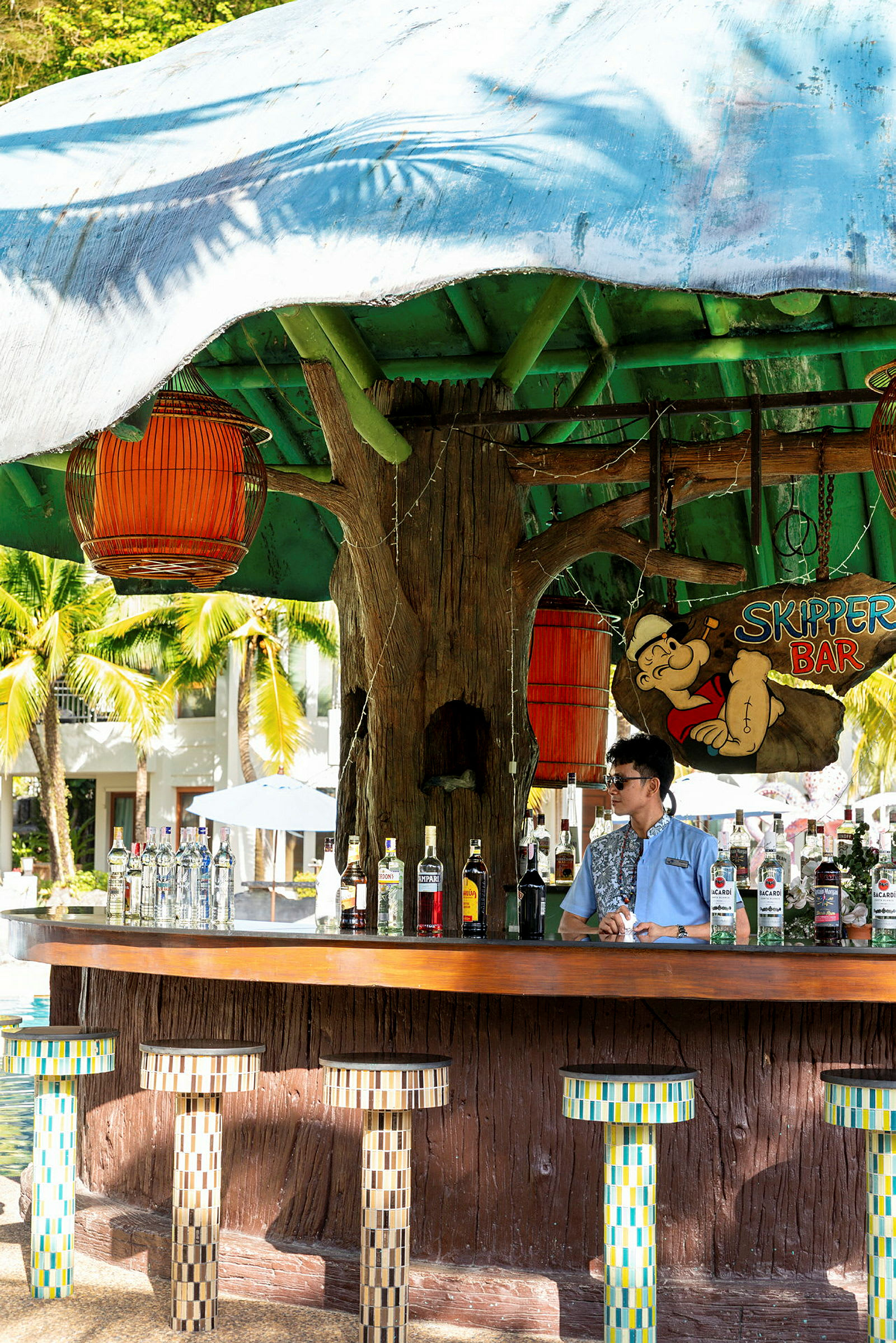 En strandbar med fargerike stoler og ulike spritflasker på bardisken. En bartender står bak baren. (AI-generert tekst)