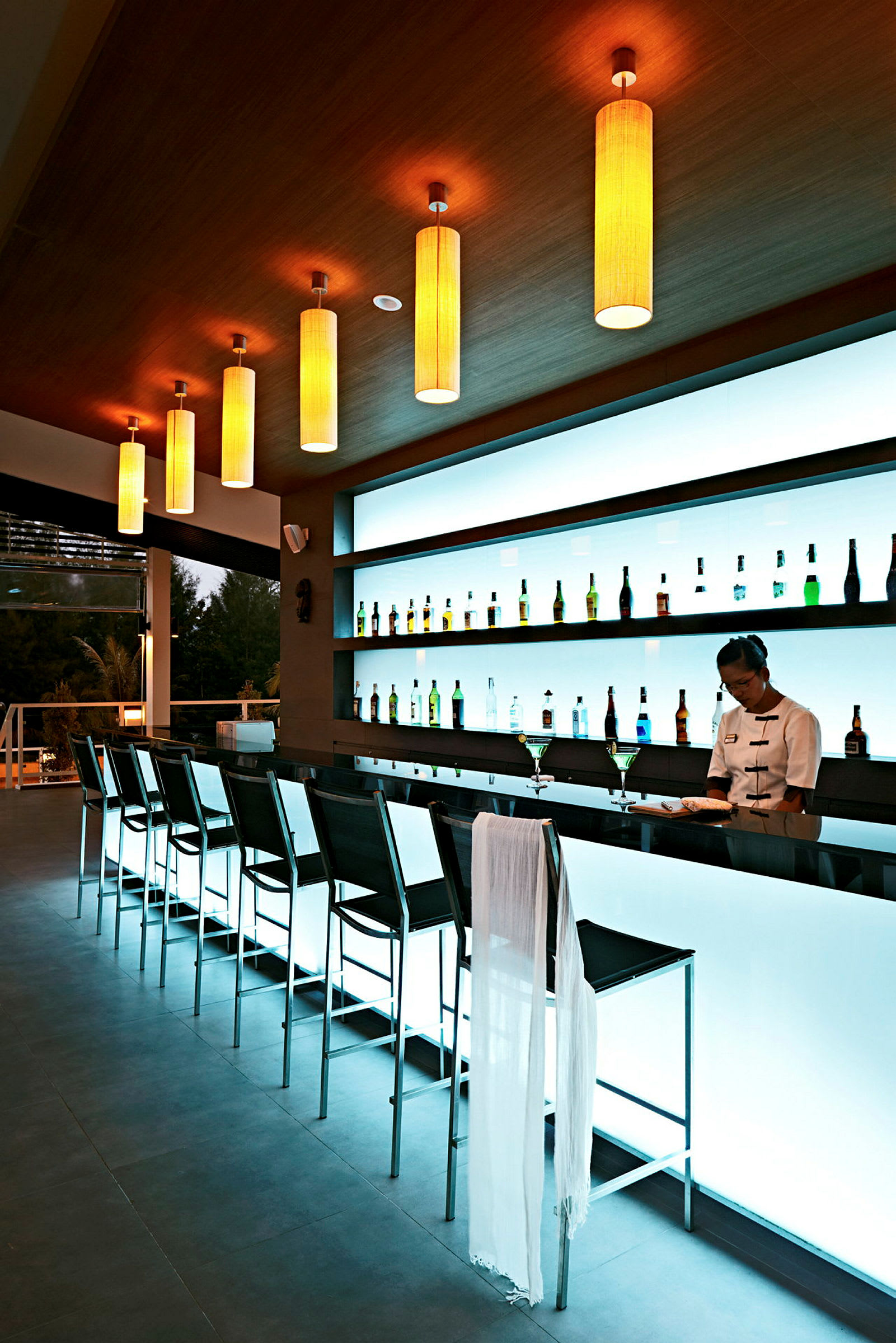Moderne bar med en rad høye stoler og opplyst bakvegg med flasker. (AI-generert tekst)