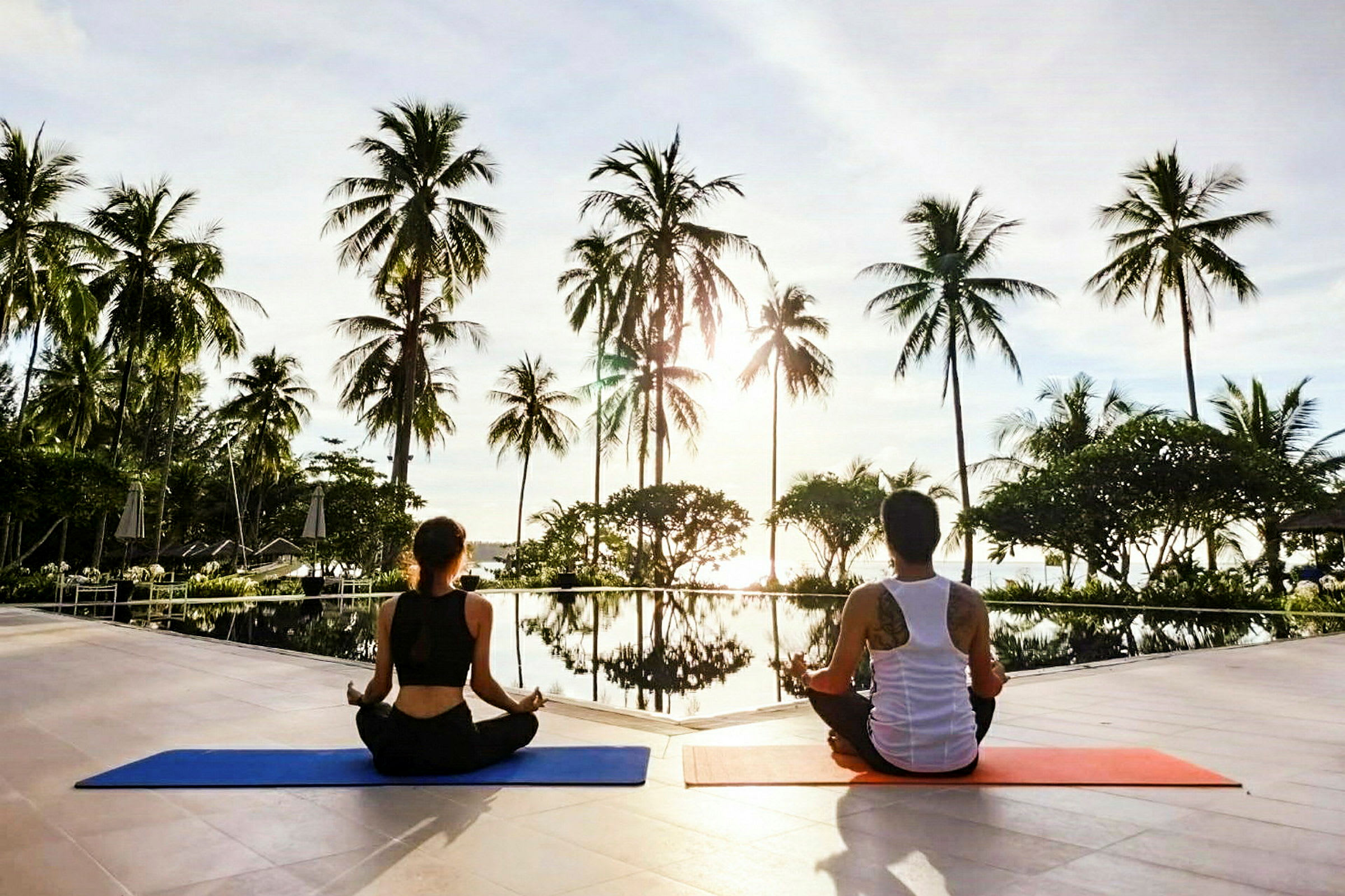 To personer sitter på yogamatter ved et basseng omgitt av palmer. (AI-generert tekst)