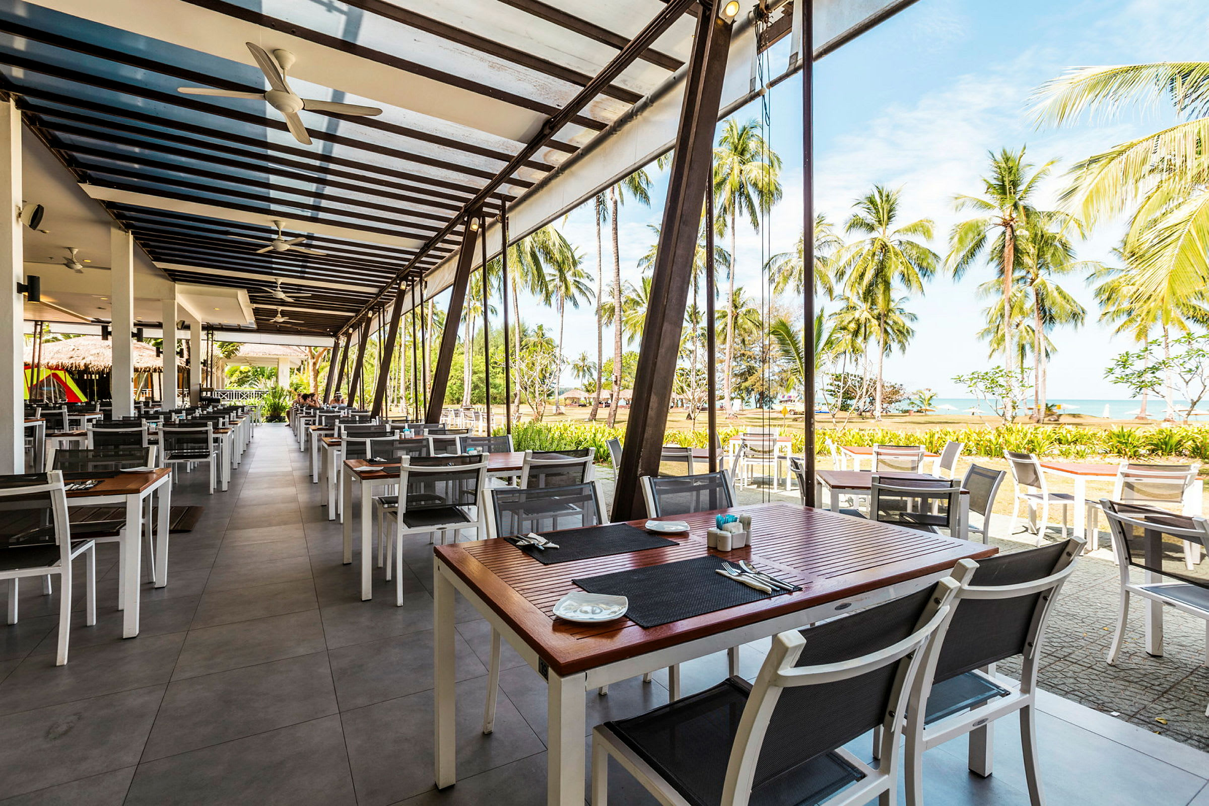 Restaurant med utemøbler under tak, med utsikt mot en strand og palmetrær. (AI-generert tekst)