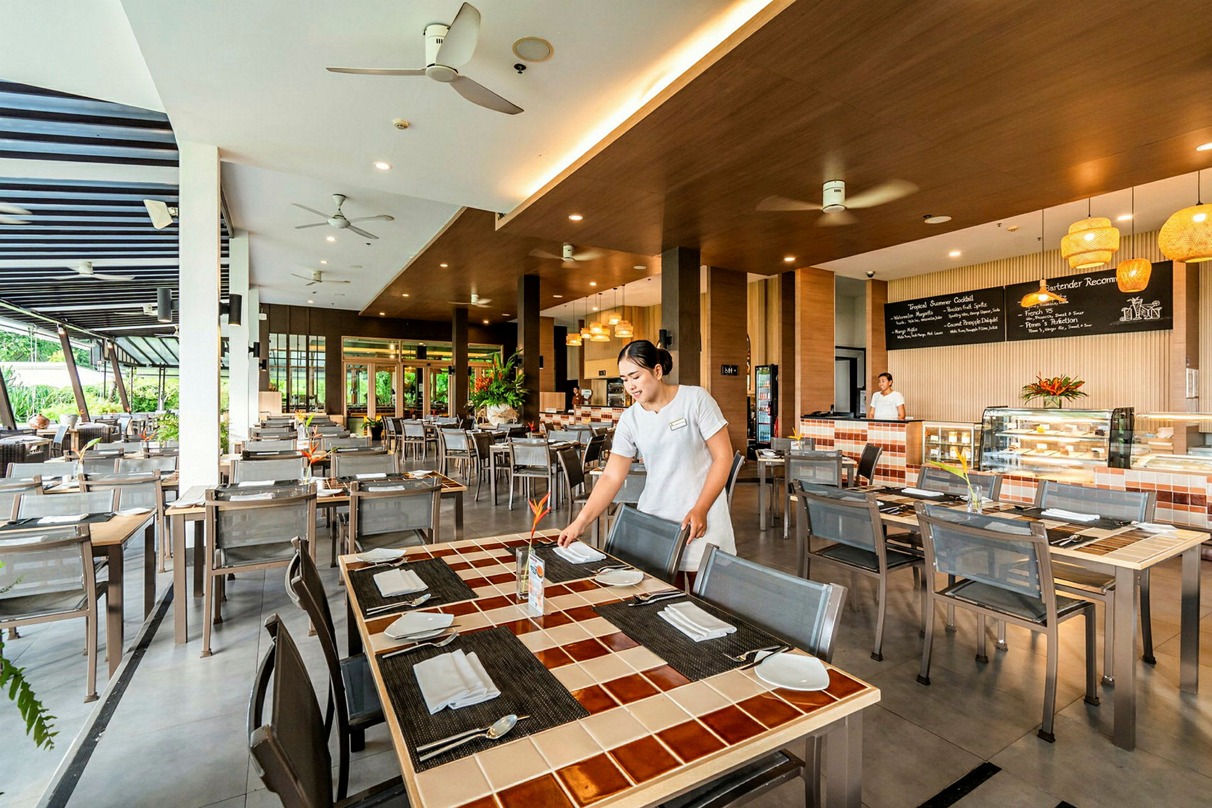 En lys restaurant med moderne møbler, en medarbeider ordner bordene. Stor bardisk i bakgrunnen. (AI-generert tekst)
