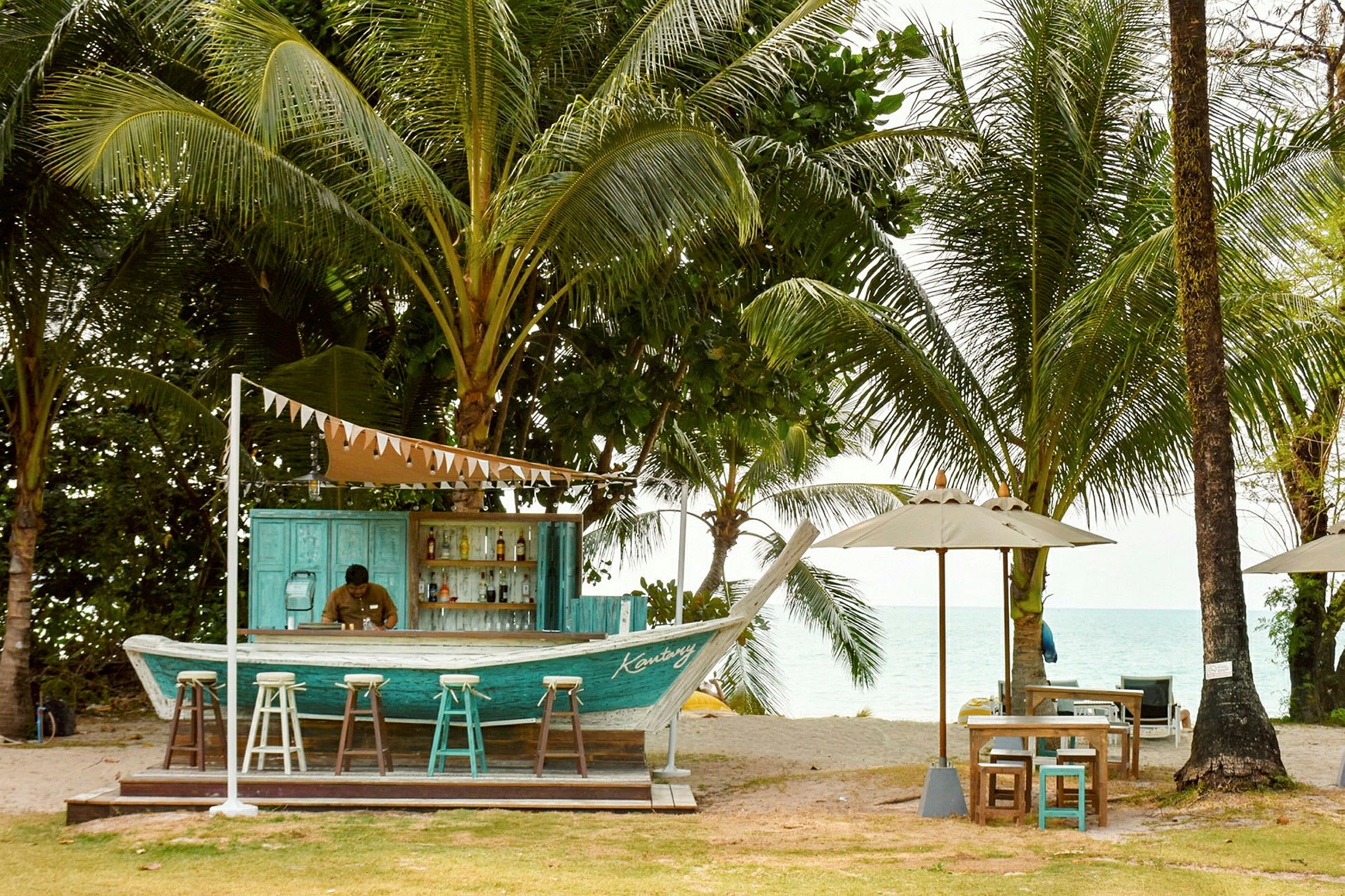 En strandbar bygget som en båt, omgitt av palmetrær og med utsikt over havet. Det er barkrakker foran baren og bord med parasoller ved siden av. (AI-generert tekst)