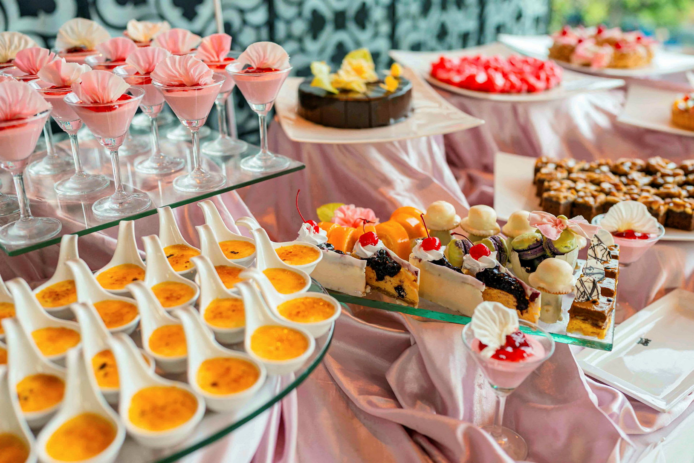 En buffet med ulike desserter, inkludert glass med rosa mousse, crème brûlée og dekorerte bakverk. (AI-generert tekst)