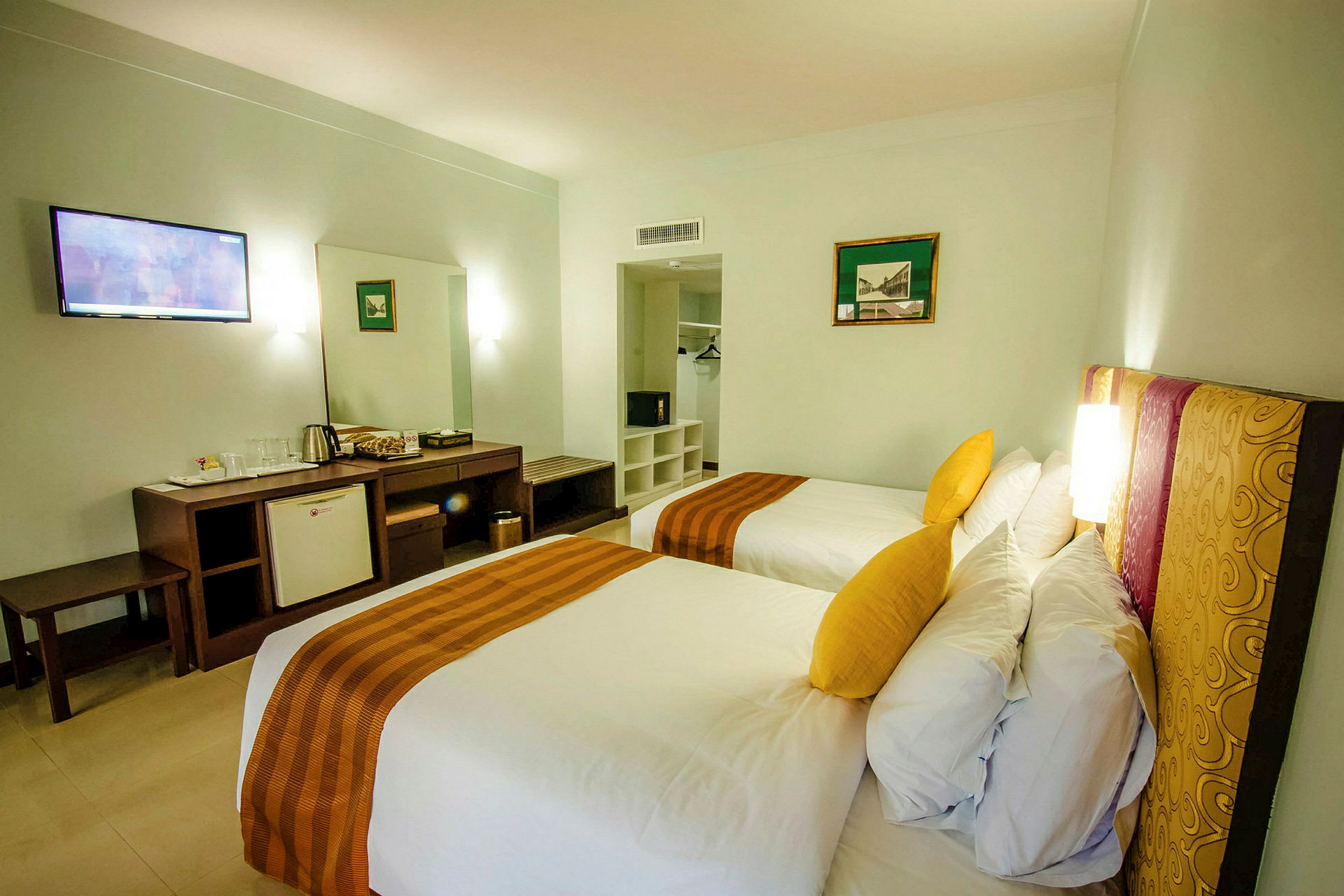 To enkeltsenger med gule puter på et hotellrom, med skrivebord, minibar og garderobe i bakgrunnen. (AI-generert tekst)