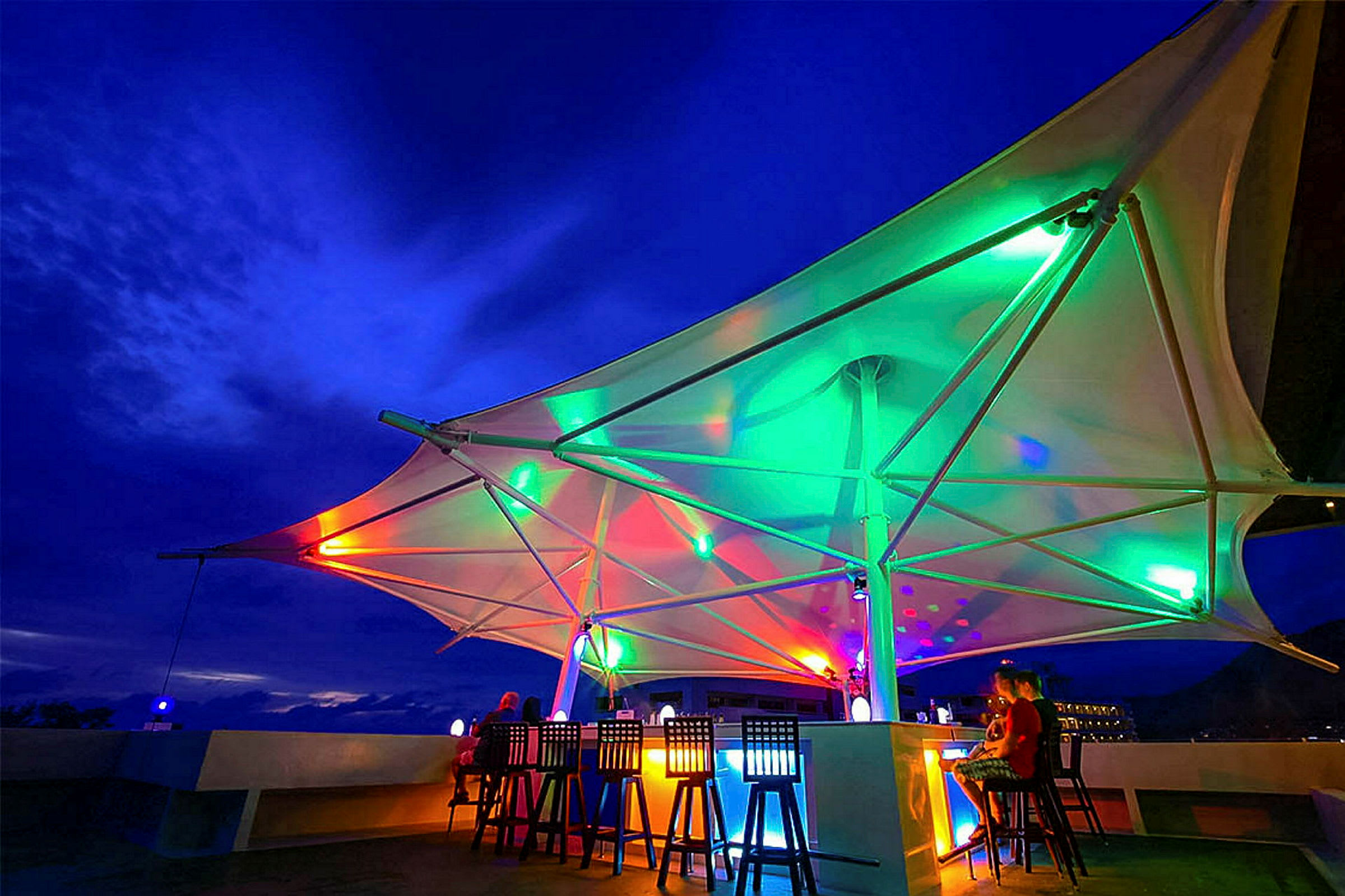 En utendørs bar med fargerik belysning og tak av seillignende materiale under nattehimmel. (AI-generert tekst)