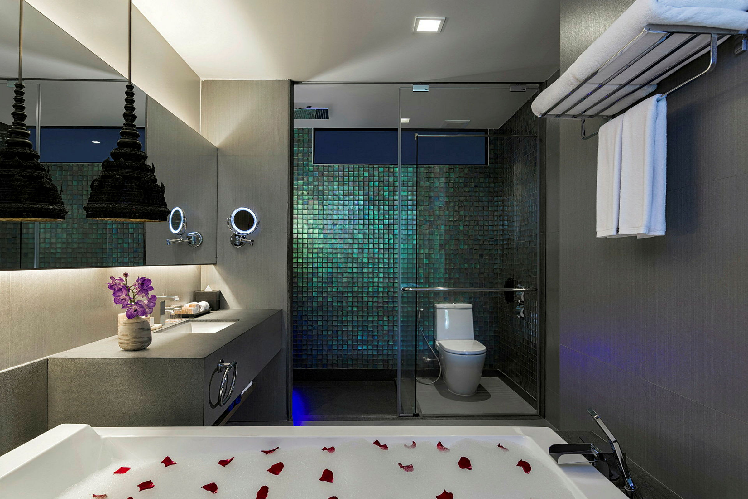 Et moderne baderom med luksuriøs innredning. Det er et badekar med skumbad og roseblader i forgrunnen. En elegant servant med speil, blomster og belysning samt en dusj og toalett bak glassvegger er synlig. (AI-generert tekst)